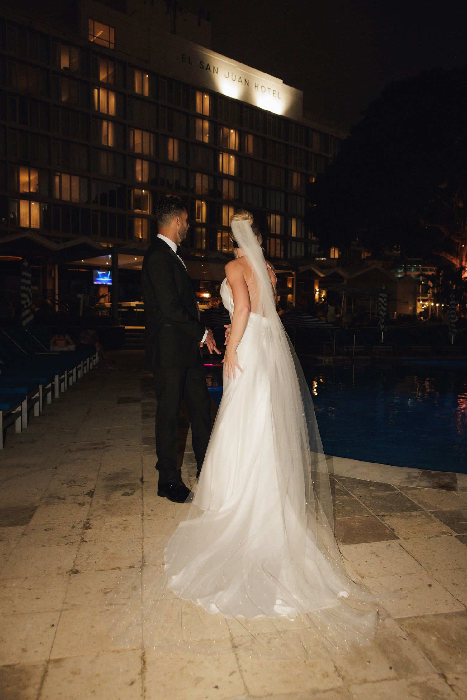 A wedding couple stands near a hotel pool at night, their silhouettes illuminated by warm building lights.