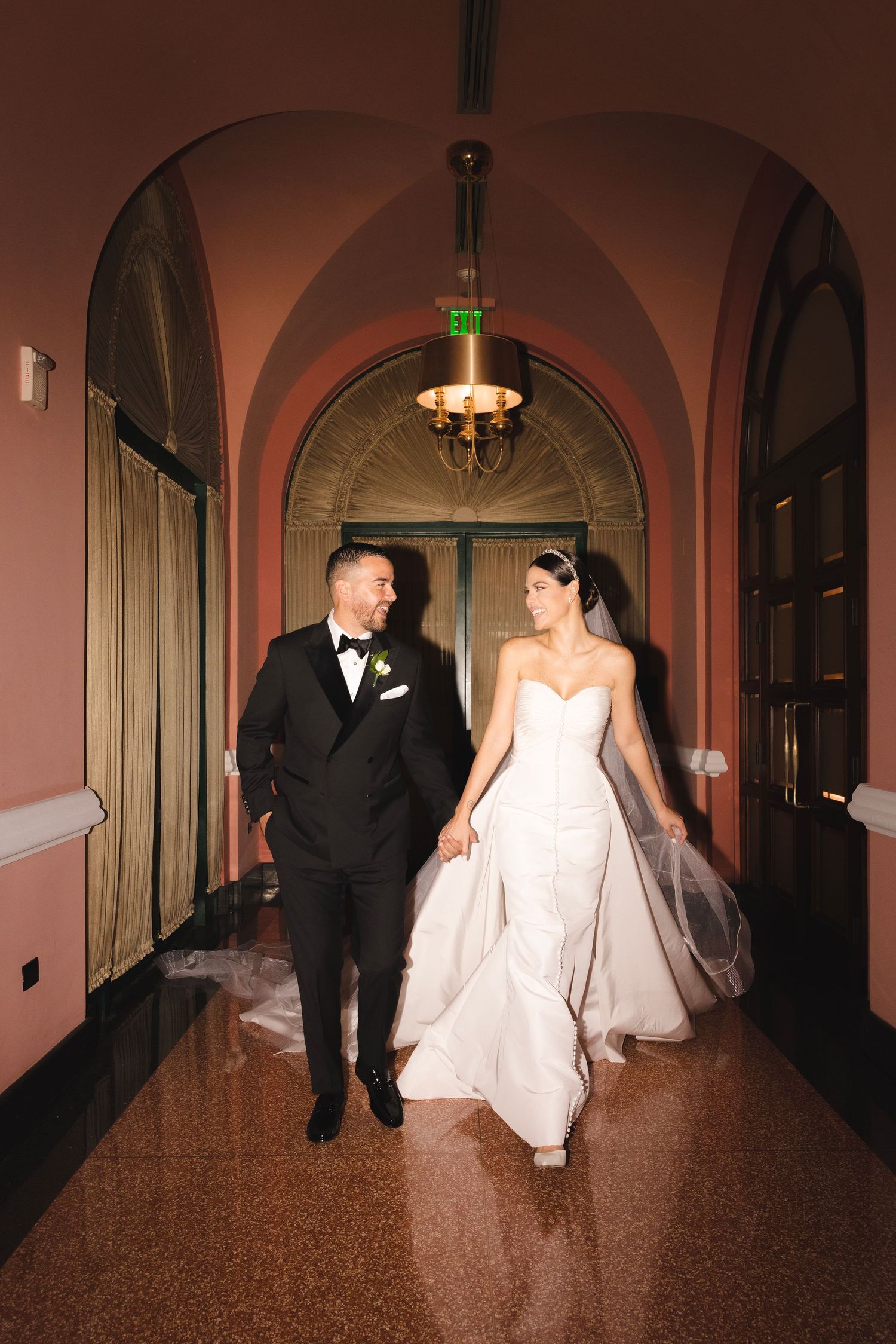 A couple walks together through an elegant arched hallway with pink walls and ornate lighting.