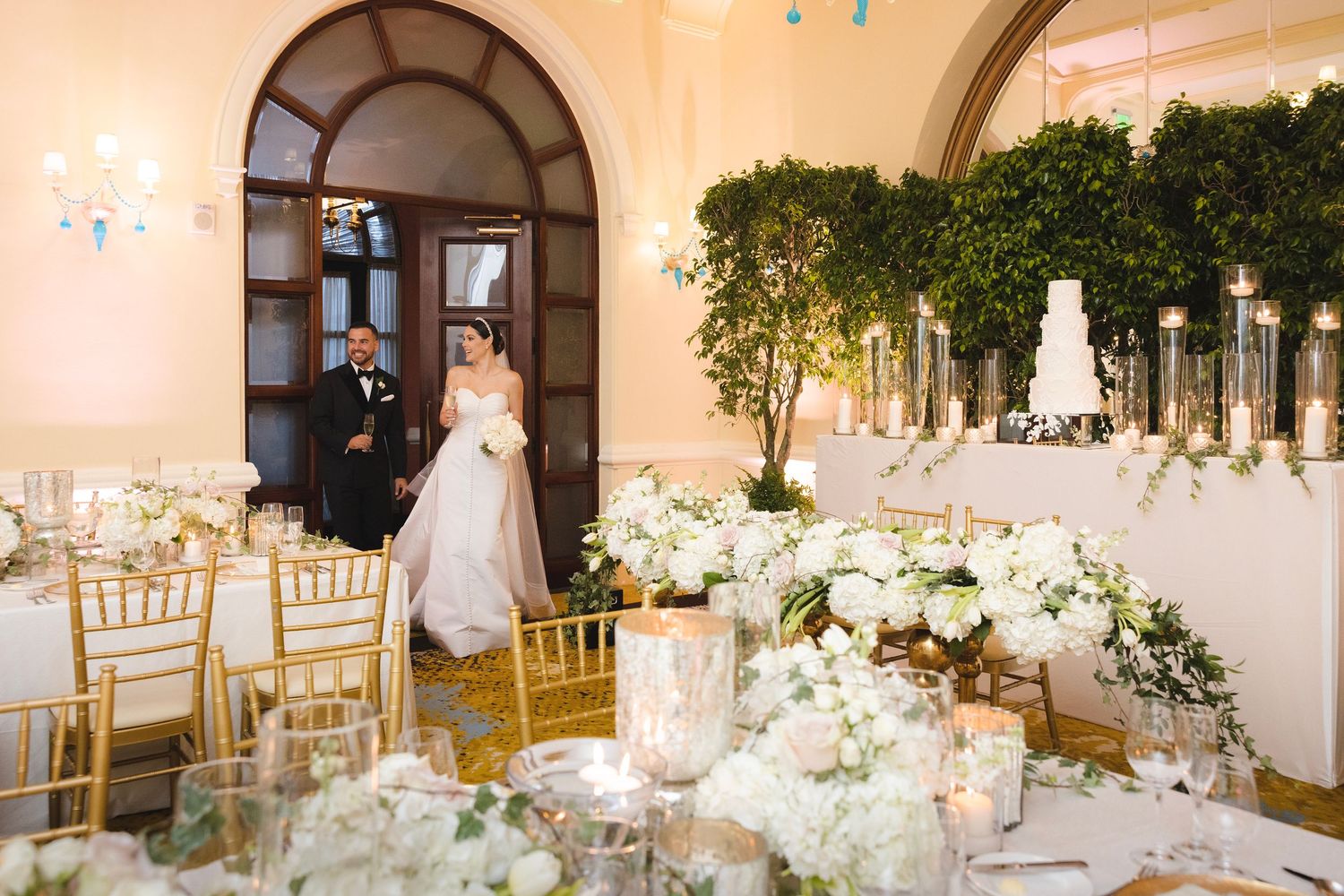 Elegant wedding reception setup with white floral centerpieces, gold chairs and candlelight in a luxurious ballroom setting.