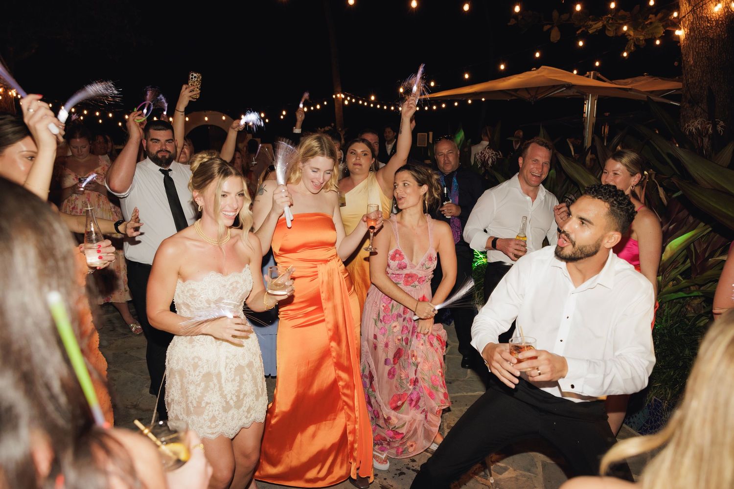 A lively dance party at an outdoor wedding reception with guests dancing under string lights in the evening.
