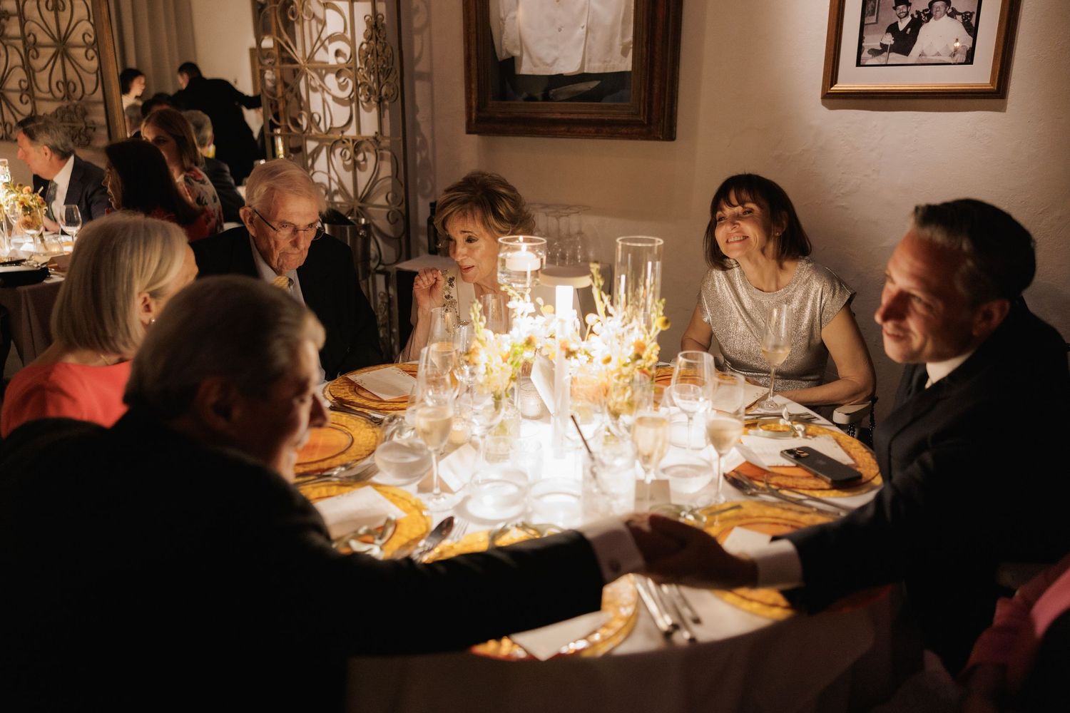 Groups of diners enjoy an elegant candlelit dinner service at multiple tables in a dimly lit restaurant setting.