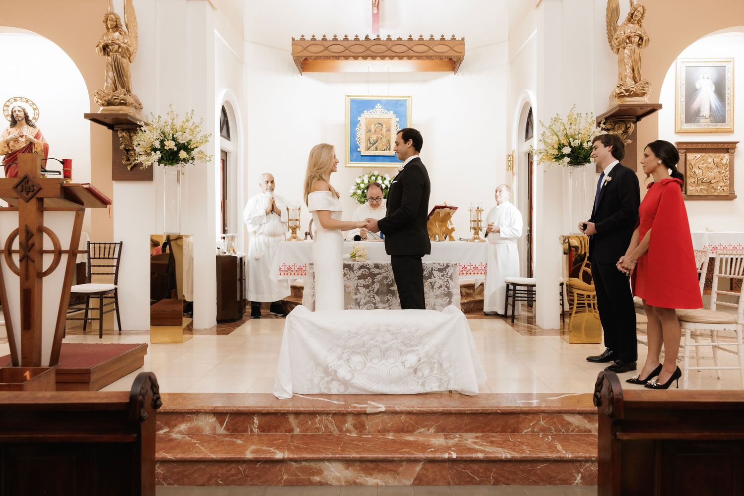 A couple stands at the altar in a white chapel with arched windows and floral arrangements during a wedding ceremony.
