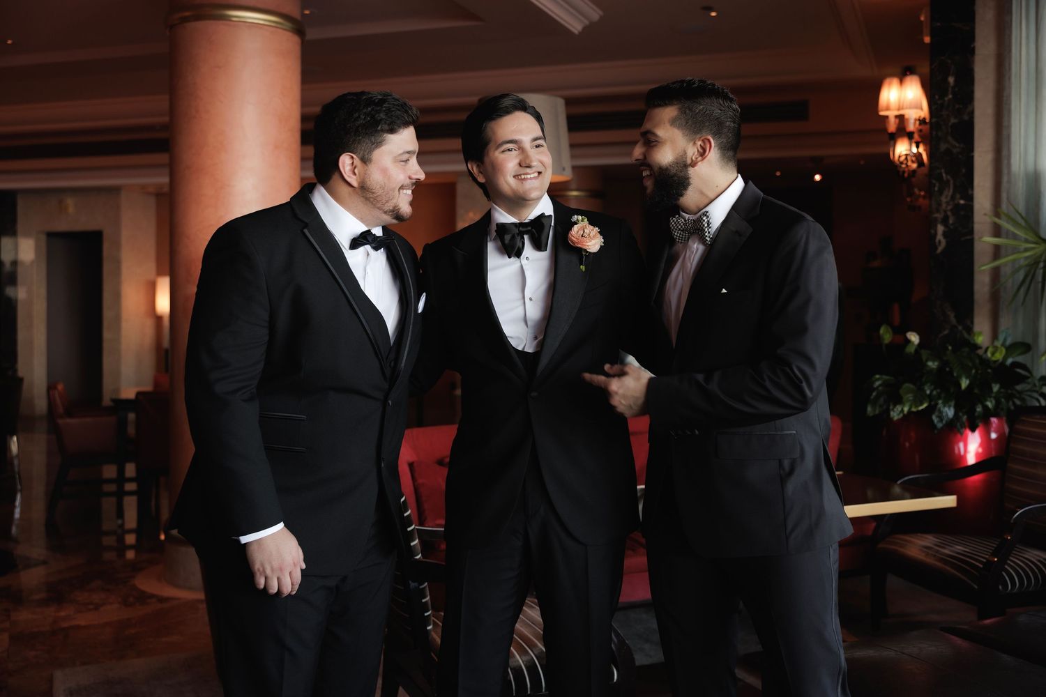 Three groomsmen in black tuxedos sharing a moment at an elegant indoor venue.