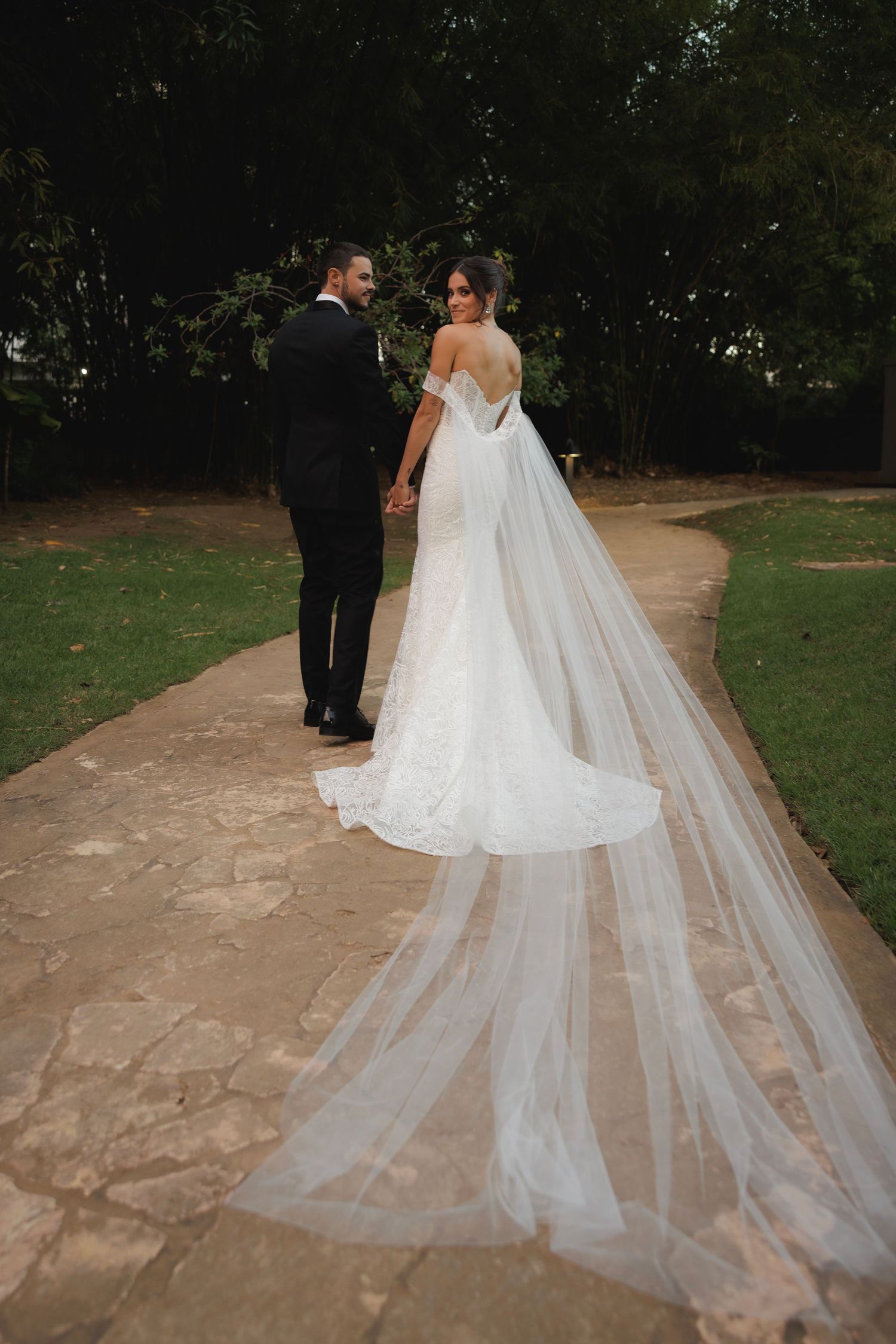Wedding couple walking down stone path with bride's flowing cathedral veil trailing behind.