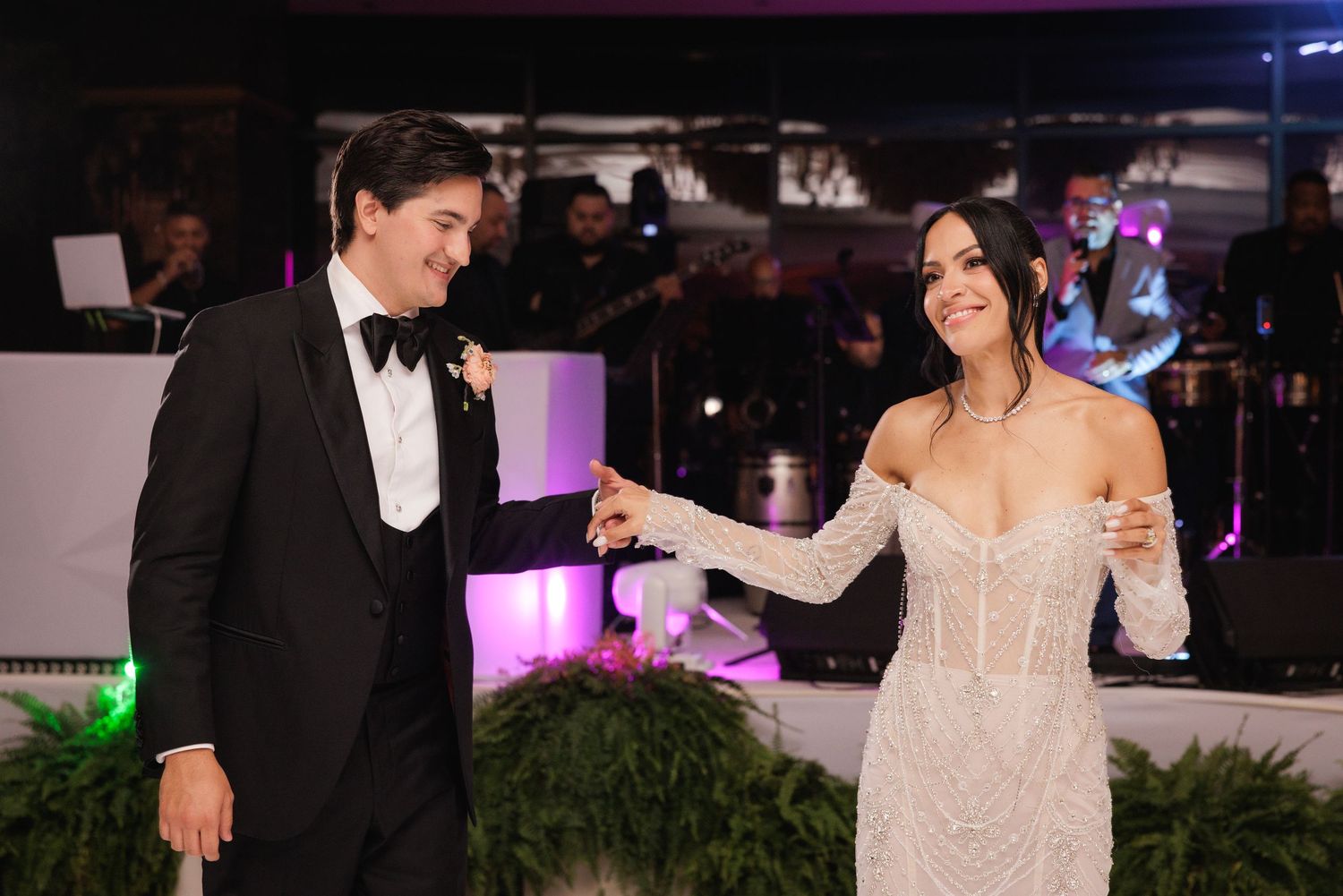 A couple in formal evening attire share a dance at a wedding reception with purple lighting and greenery decor.