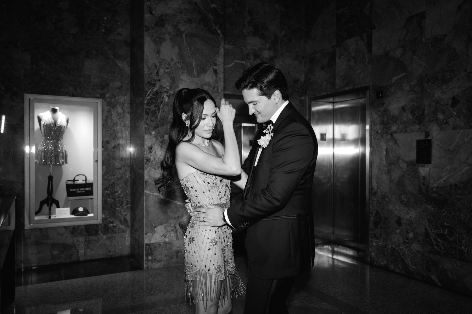 A couple in formal evening wear share an intimate moment in an elegant hallway with marble walls and dim lighting.