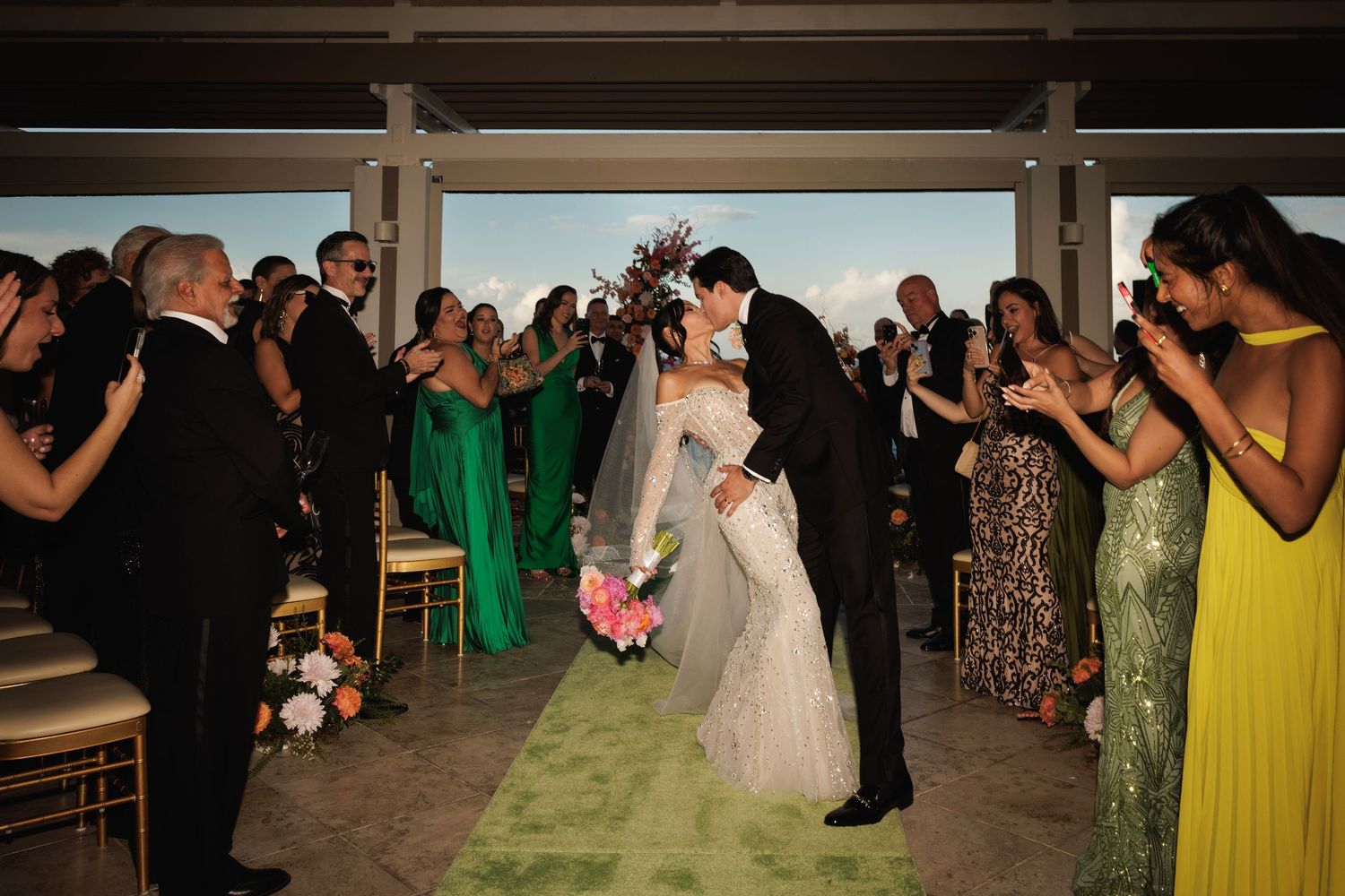 Wedding guests line both sides of a green carpet aisle as they photograph a couple in a traditional ceremony.