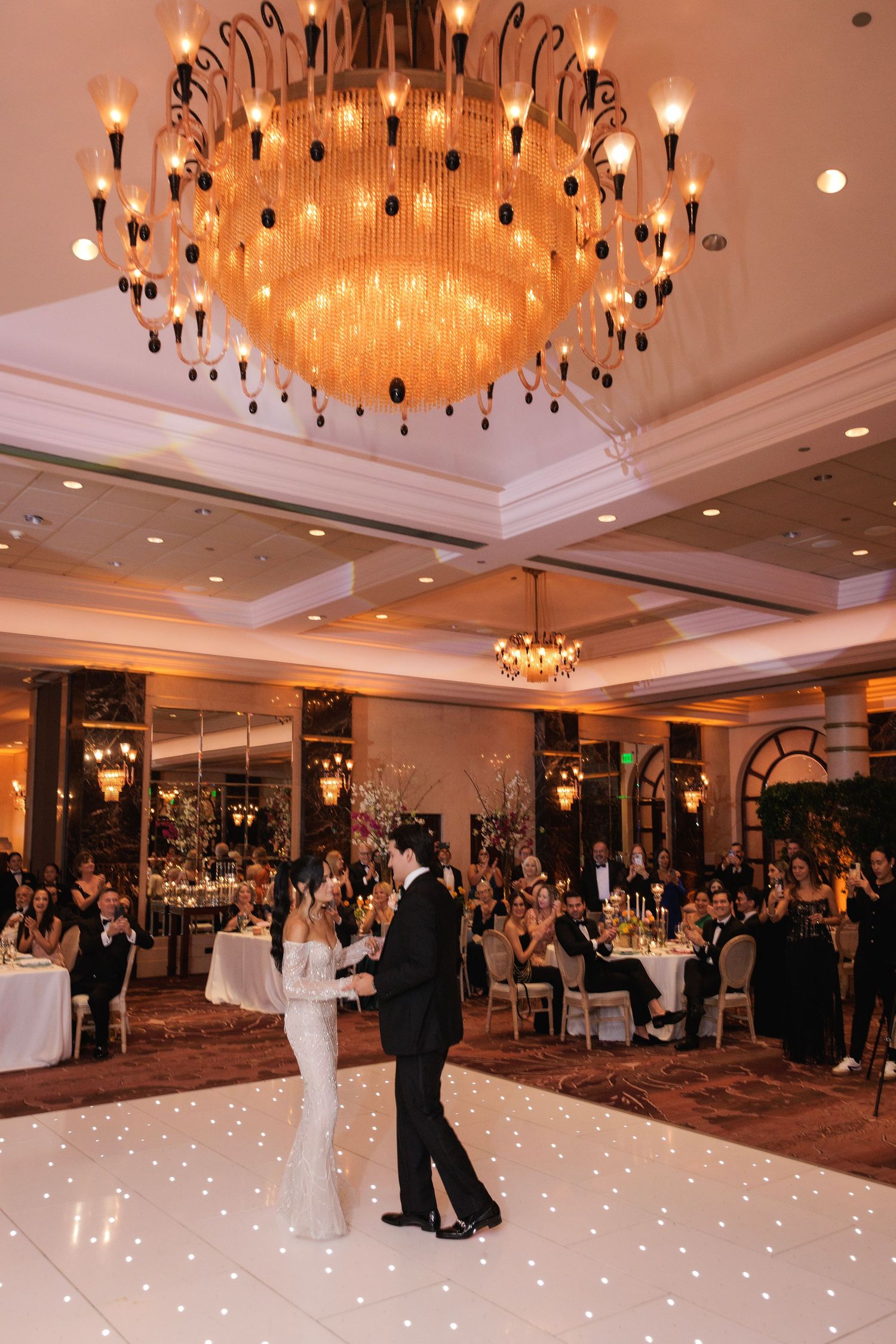 A couple shares their first dance on an illuminated white dance floor under a grand crystal chandelier in an elegant ballroom.