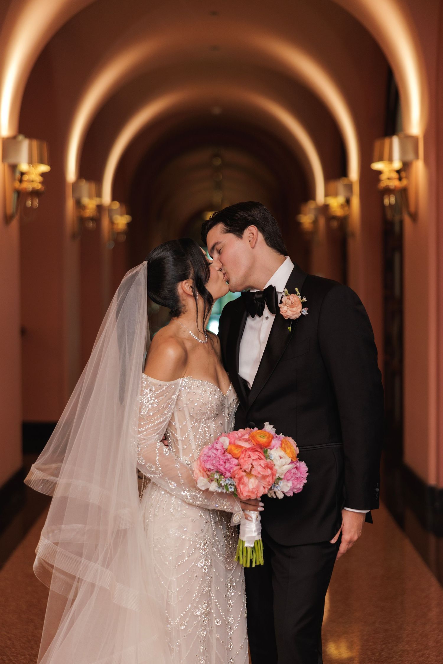 Newlyweds share an intimate moment in an elegant hallway with arched ceilings and wall sconces.
