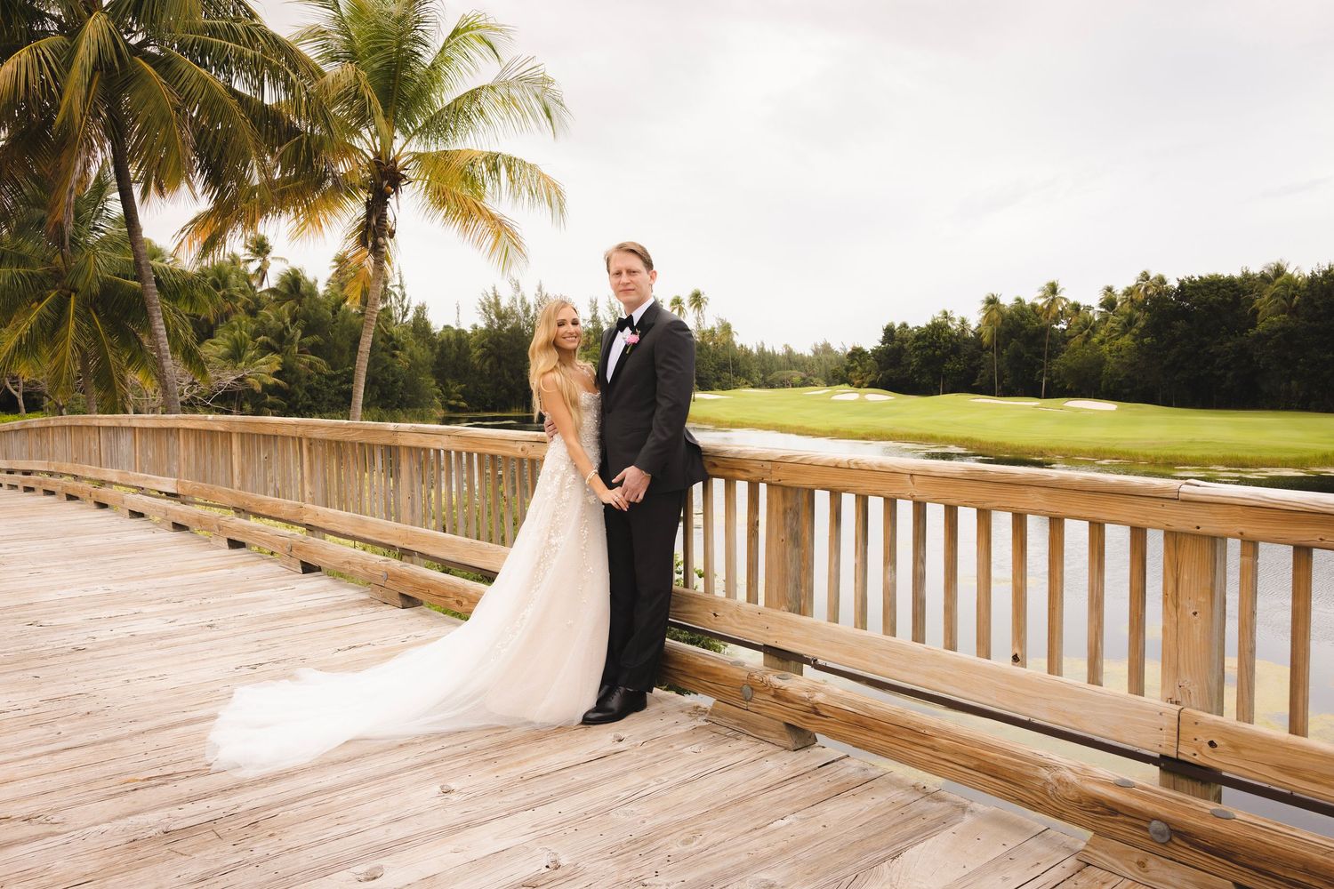 A wedding couple stands on a wooden boardwalk overlooking a tropical golf course with palm trees at sunset.