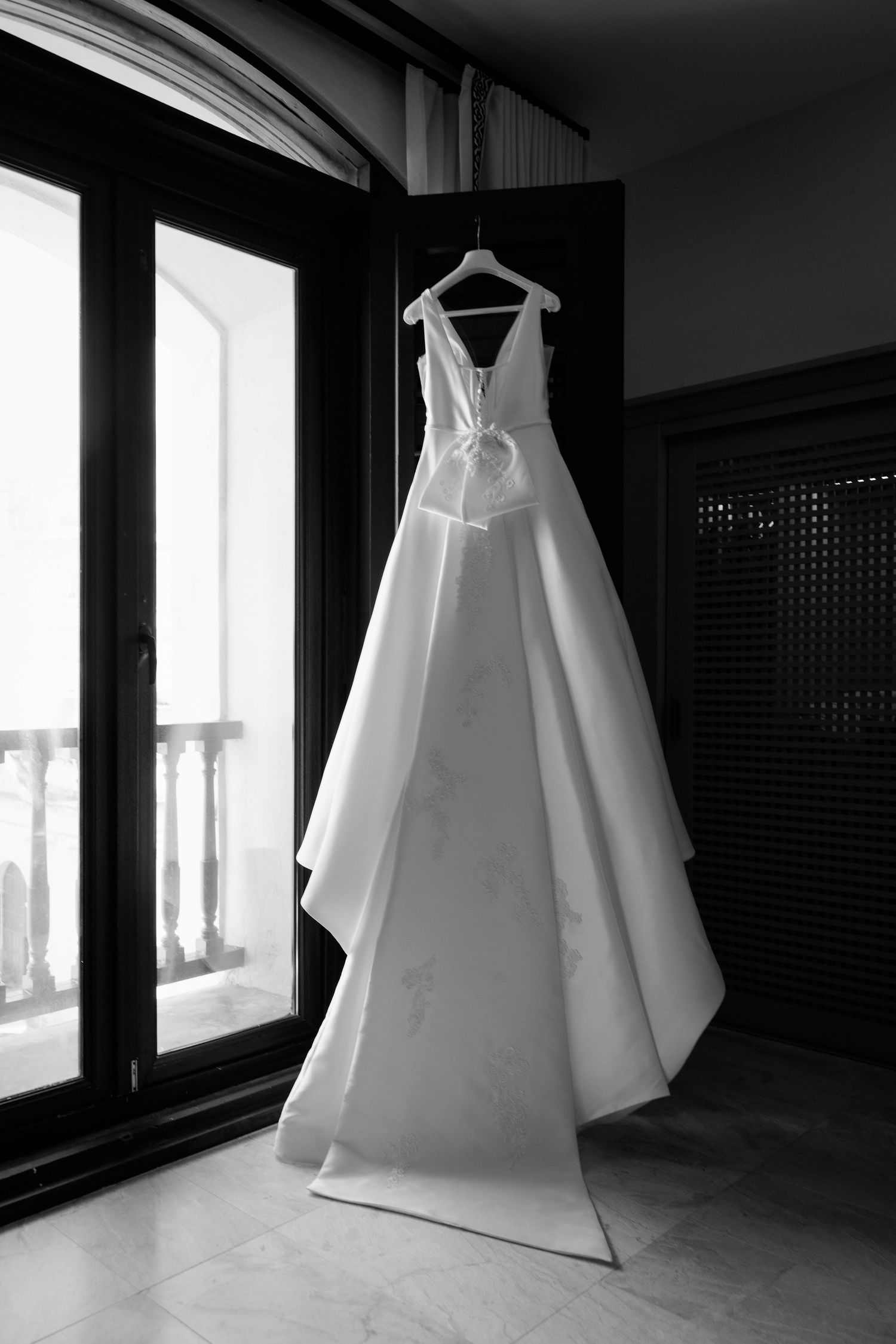 Elegant white wedding dress hanging in front of large windows in a dark room.