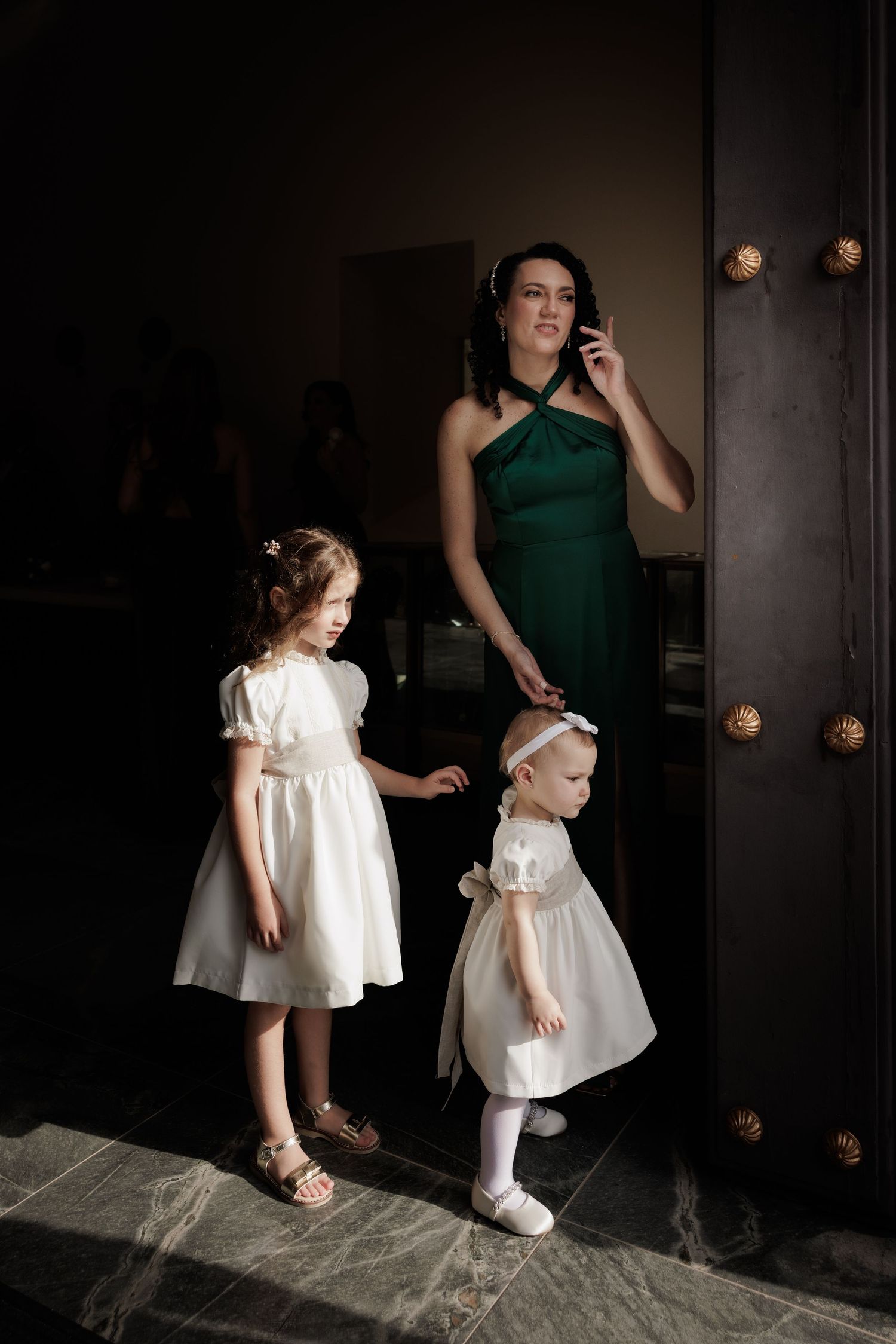 A child in a white dress stands near a dark doorway while being helped by someone in green.