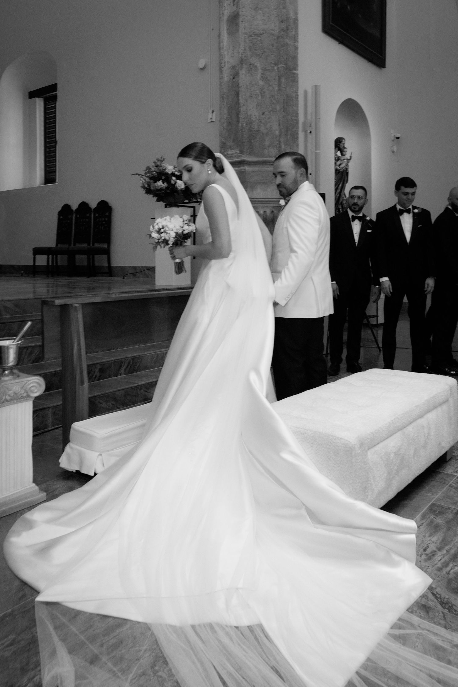 A wedding ceremony in a church with a flowing white bridal gown and groom in white tuxedo jacket.