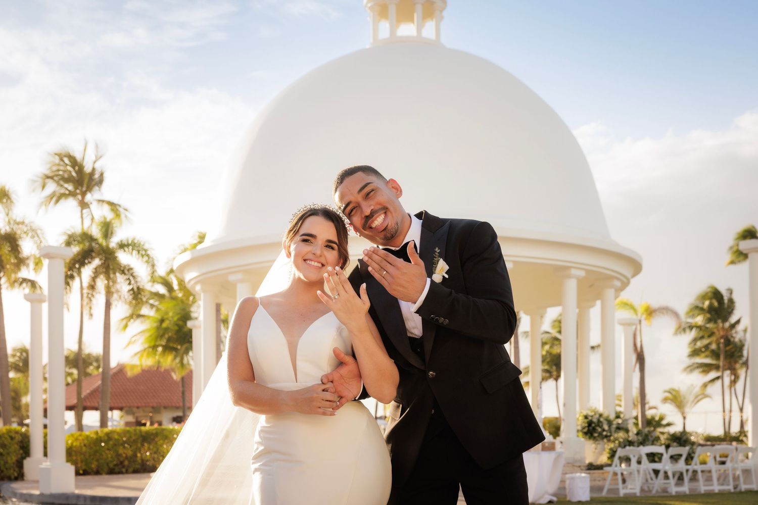 Happy couple shares tender moments during sunset beach ceremony with palm trees and white gazebo in background.