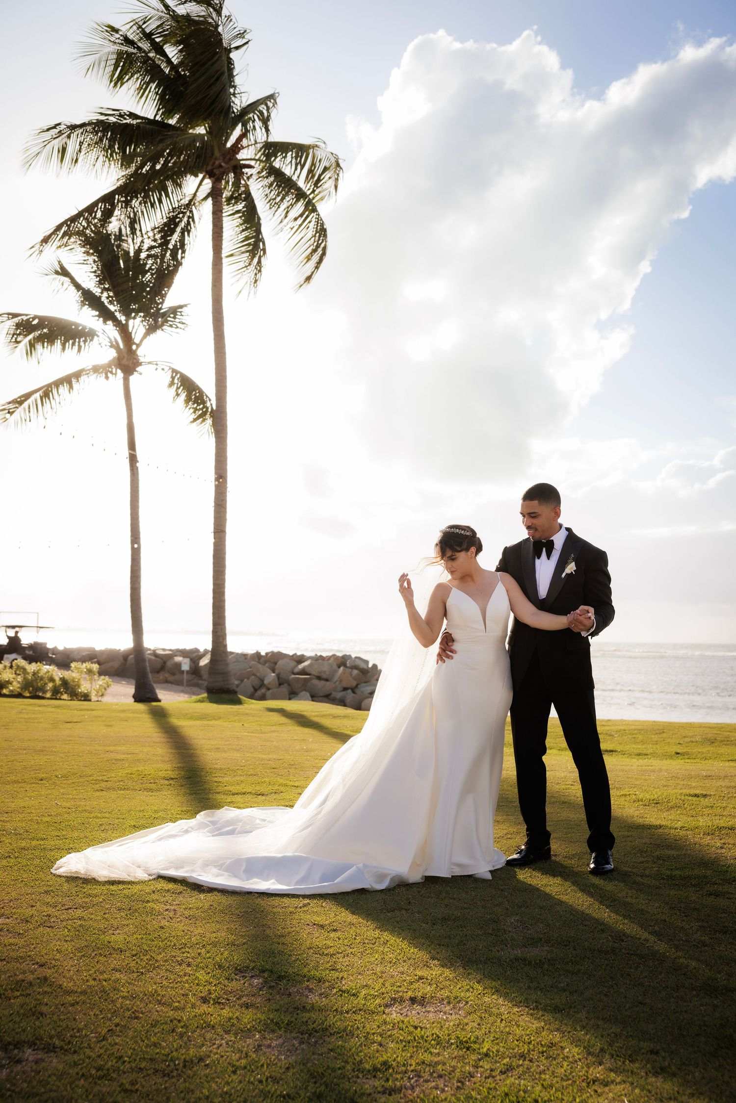 Wedding couple walks together on grassy lawn with palm trees and ocean view at sunset in tropical paradise.