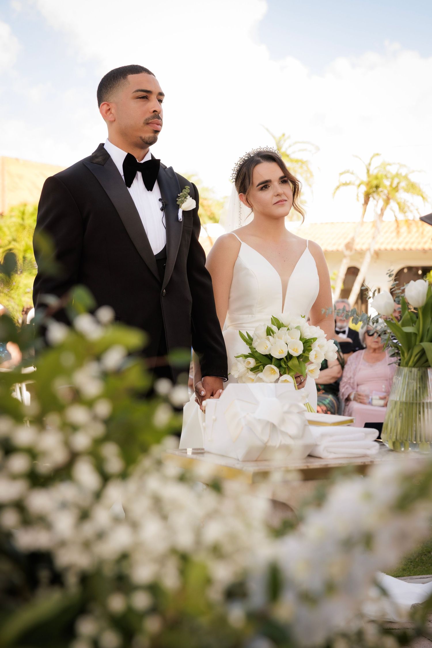 An elegant wedding couple sits at their reception table decorated with white floral arrangements and blurred cherry blossoms.
