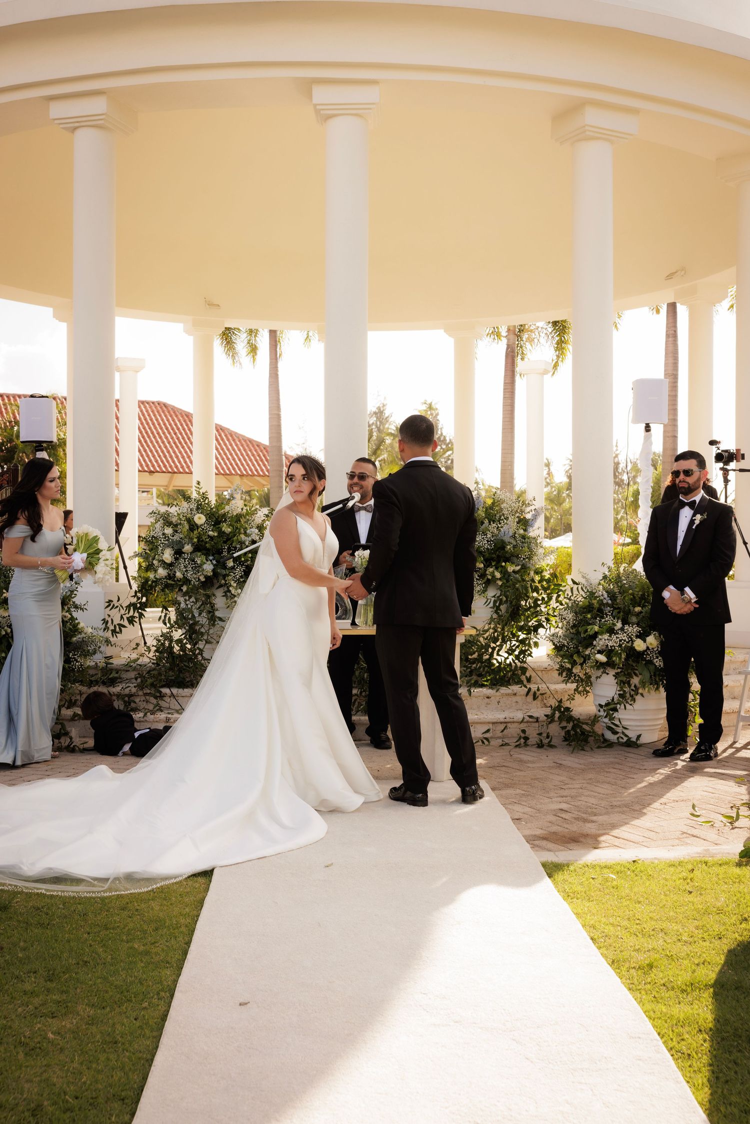 Elegant outdoor wedding ceremony at a columned venue with white pillars and flowing dress train in golden sunlight.