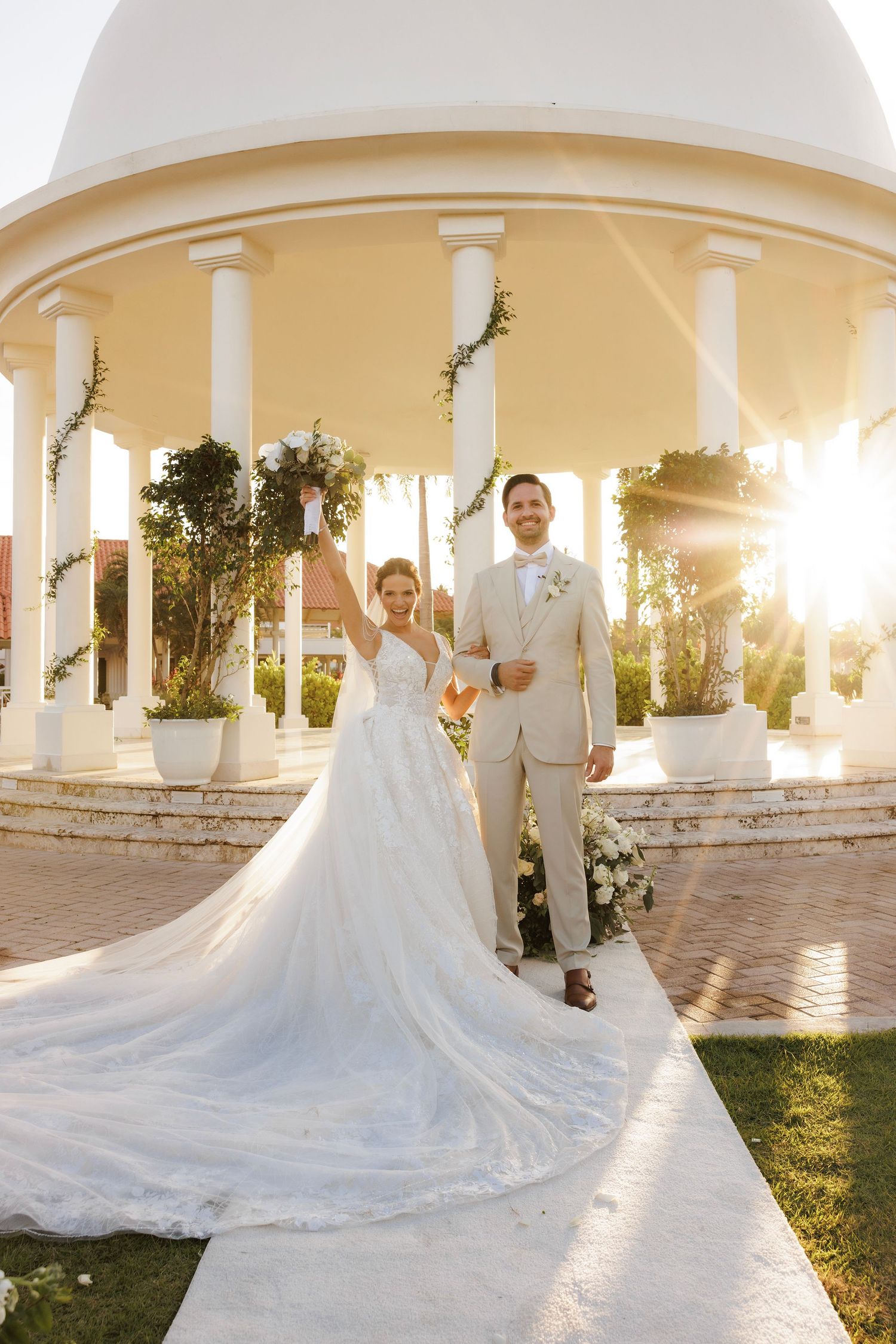 Elegant outdoor wedding ceremony at sunset with white pillars and a flowing wedding dress train sweeping across the courtyard.