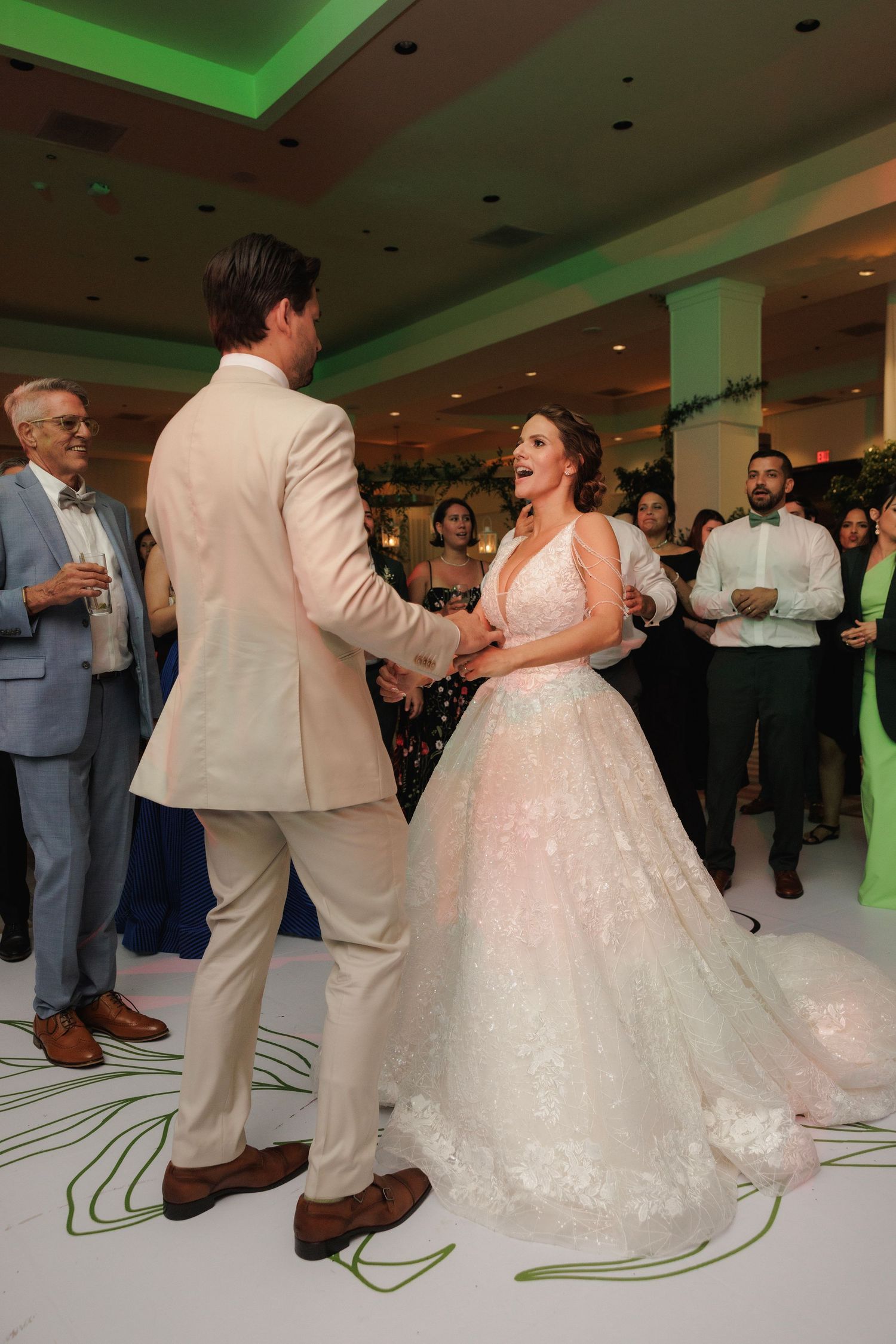 Wedding couple performs their first dance surrounded by guests in a reception hall with green accent lighting.