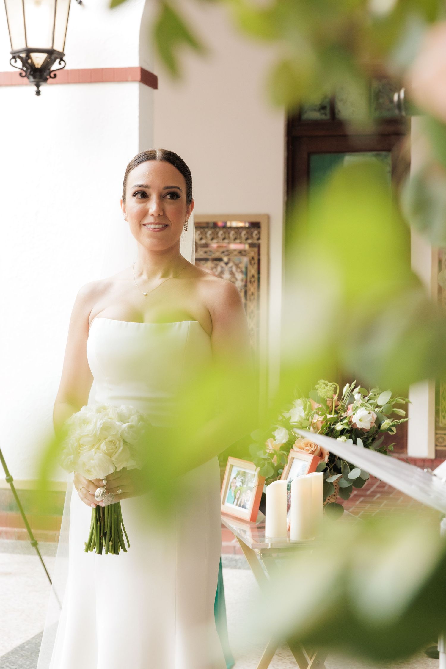 A bride in a sleek white strapless gown holds a bouquet of white flowers in an elegant indoor setting.