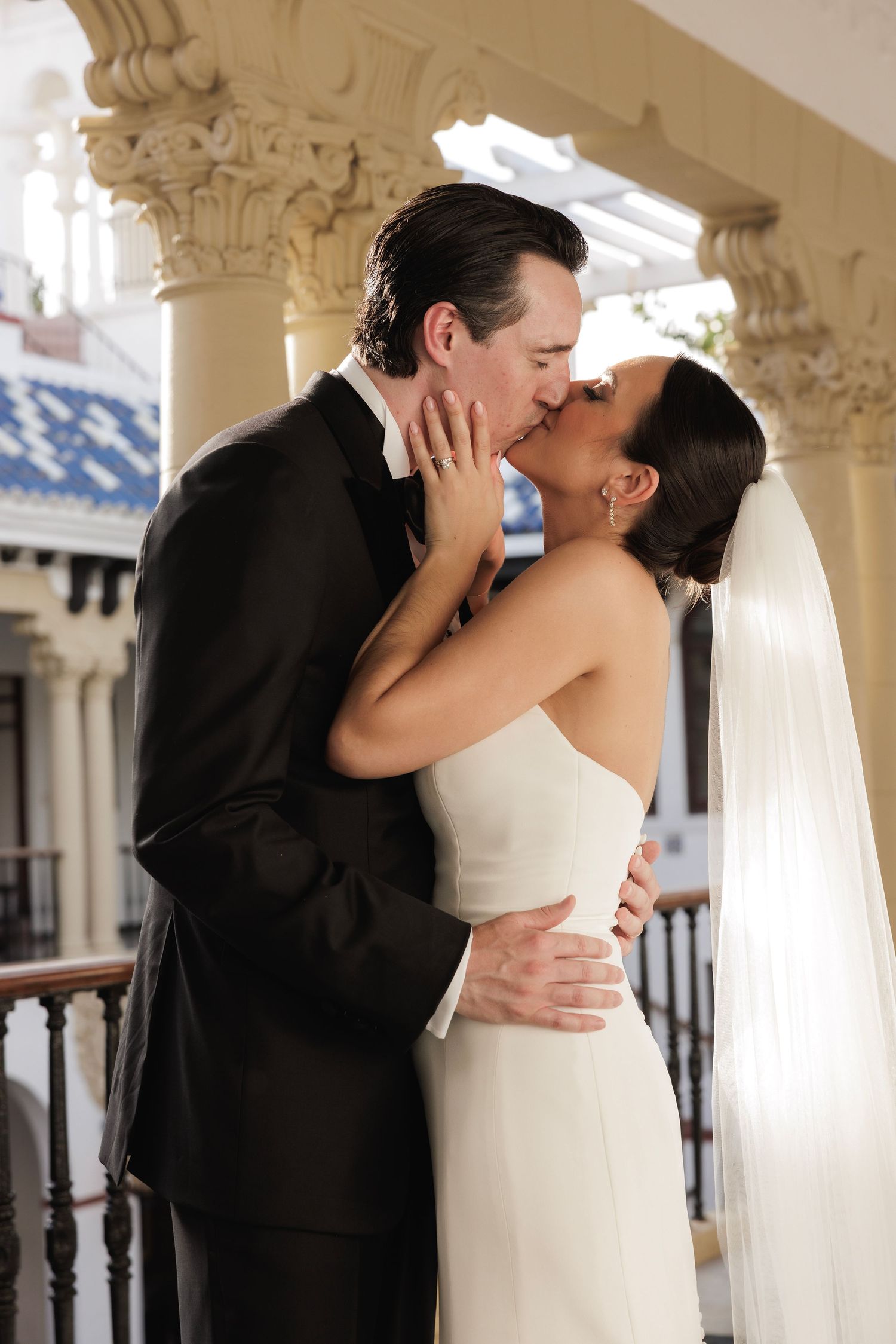 Newlywed couple sharing a romantic kiss on an elegant balcony with ornate architectural columns in the background.