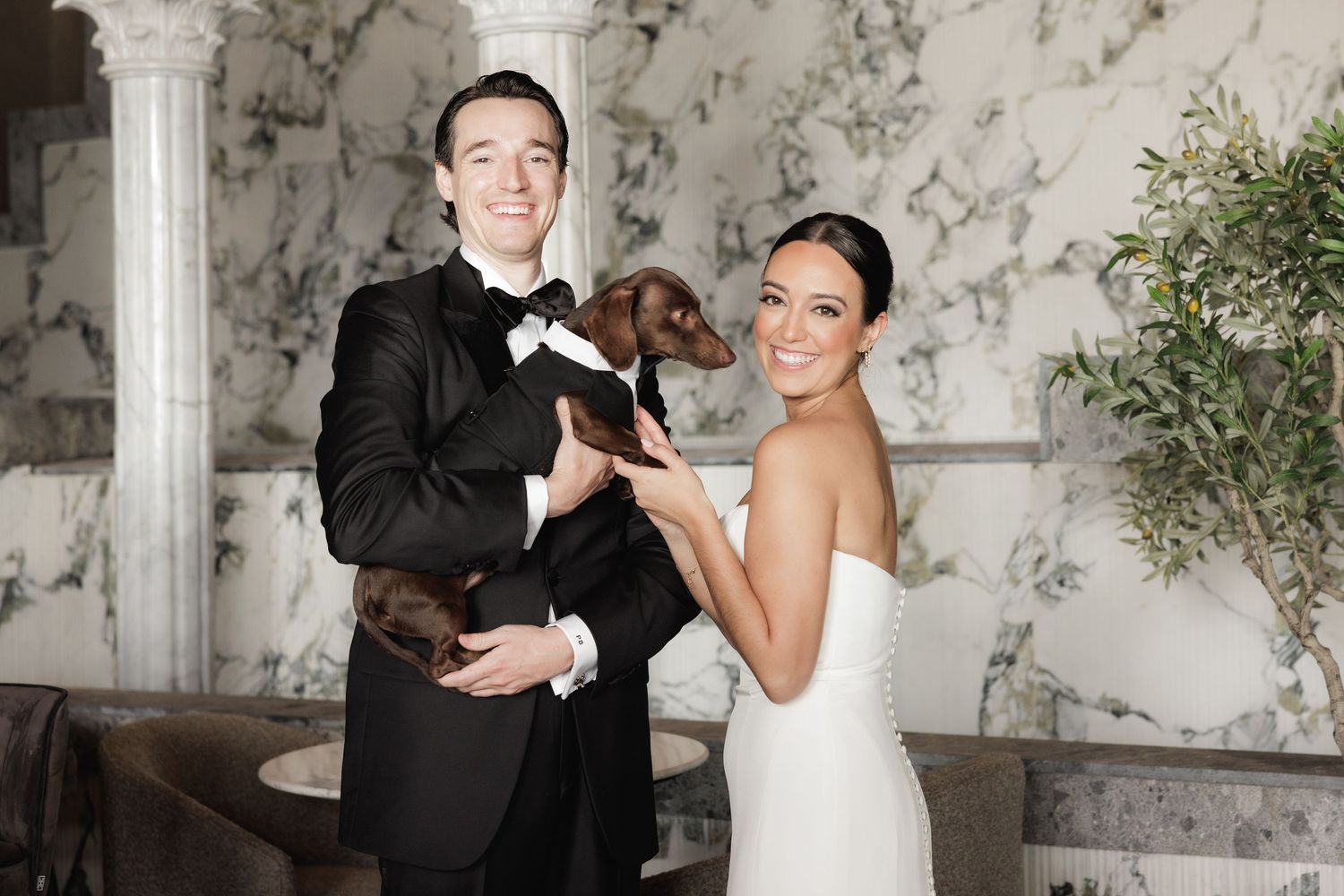 Bride and groom share joyful moments with their pet dog during elegant wedding photos in marble-walled venue.