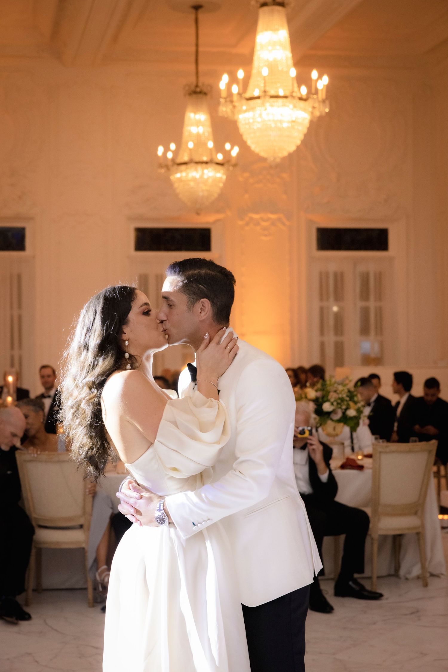 A couple shares an intimate dance moment under warm chandelier lighting in an ornate ballroom.