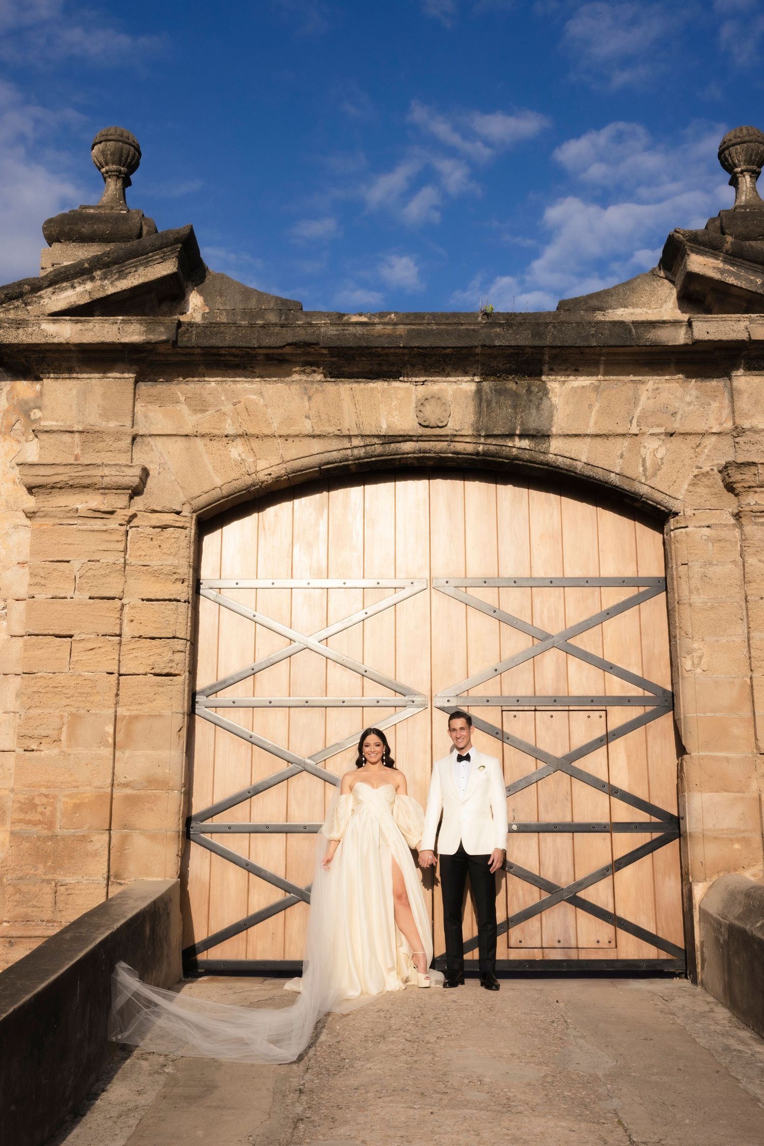 Wedding couple poses against rustic stone wall with large wooden gates in series of shots.