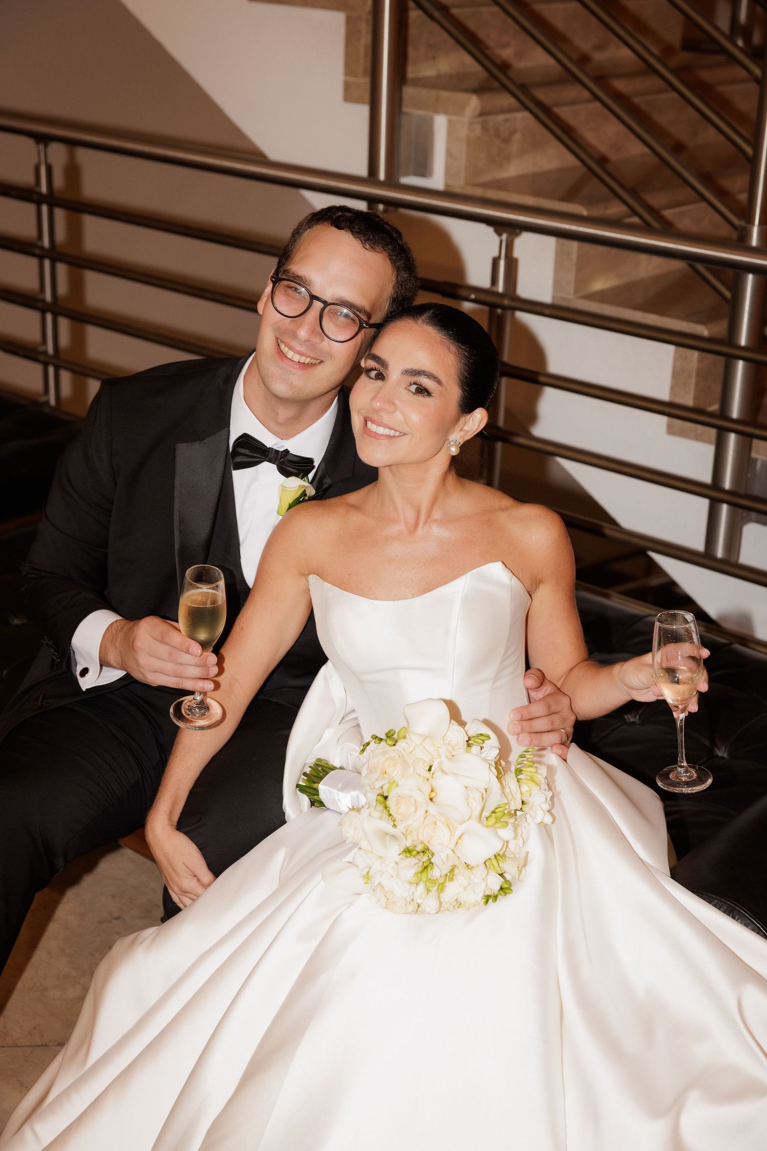 A newlywed couple celebrates with champagne while sitting on modern stairs.