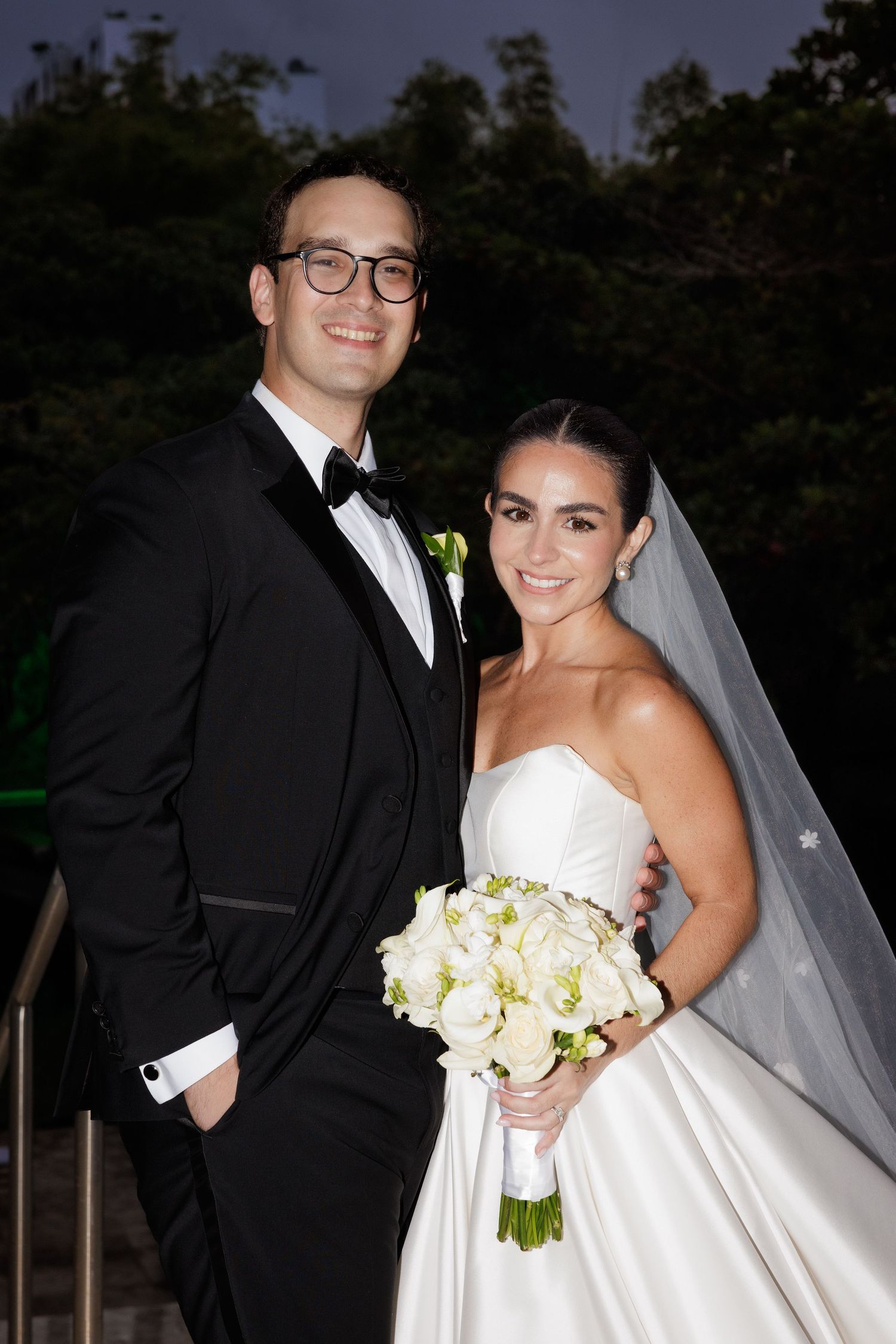 Evening wedding portraits showing a couple in formal attire with white bouquet against dark sky.
