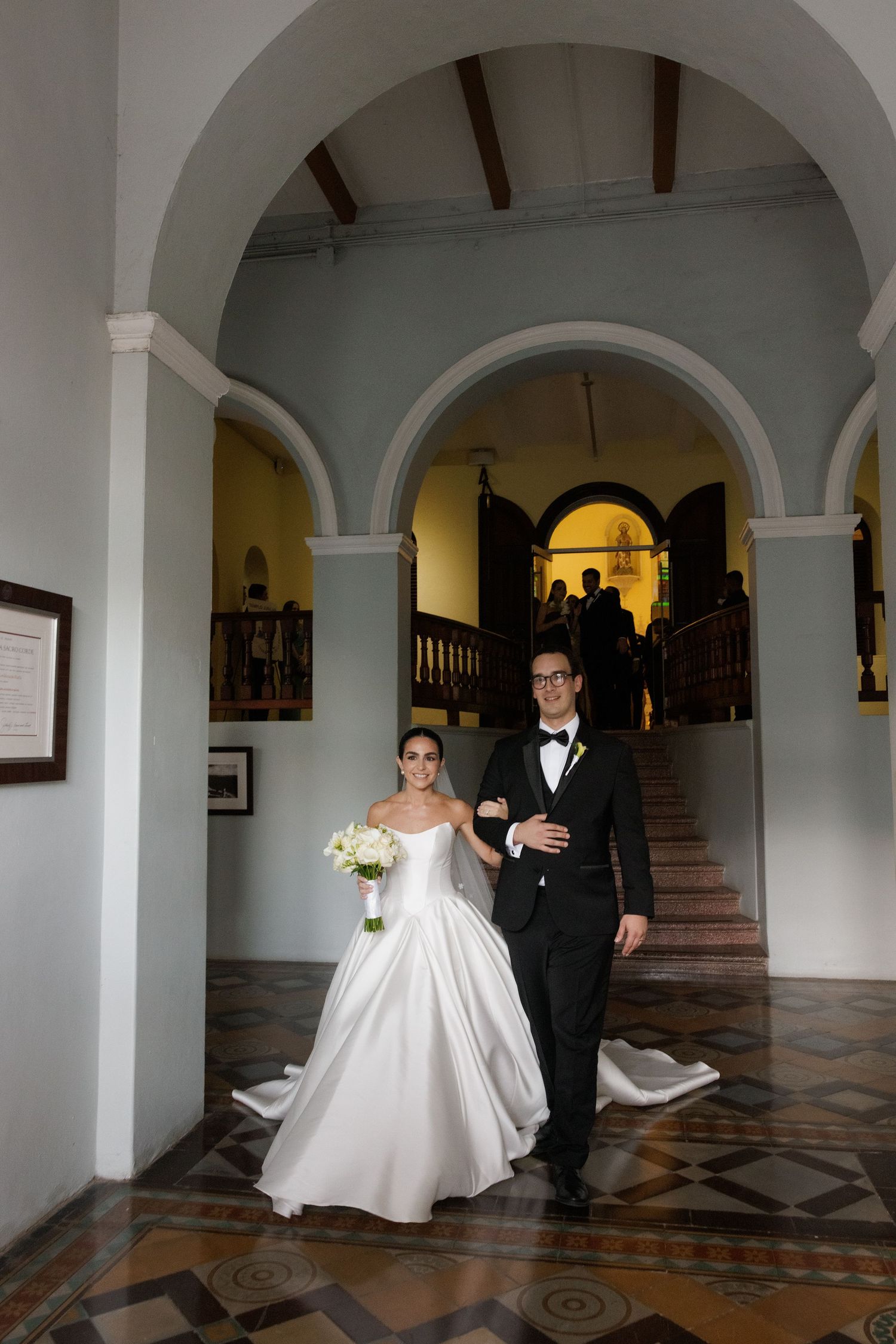 Elegant couple walks through arched hallway with checkerboard floor and cream-colored walls.