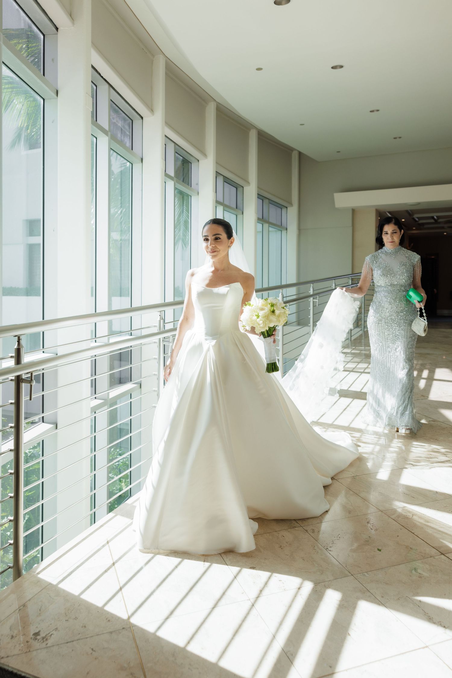 Elegant bridal gowns displayed in sunlit hallway with floor-to-ceiling windows and modern railing.