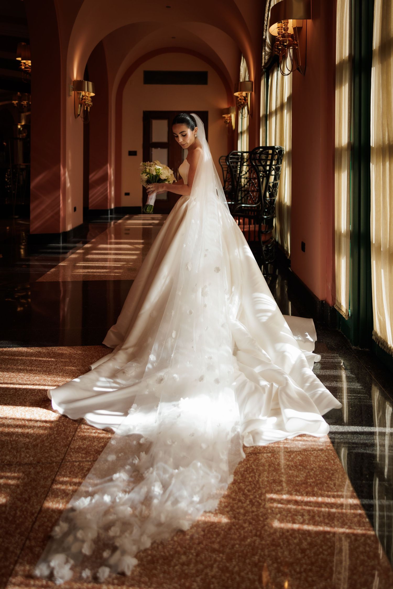 A bride in a flowing white gown stands in an elegant hallway with dramatic lighting streaming through windows.