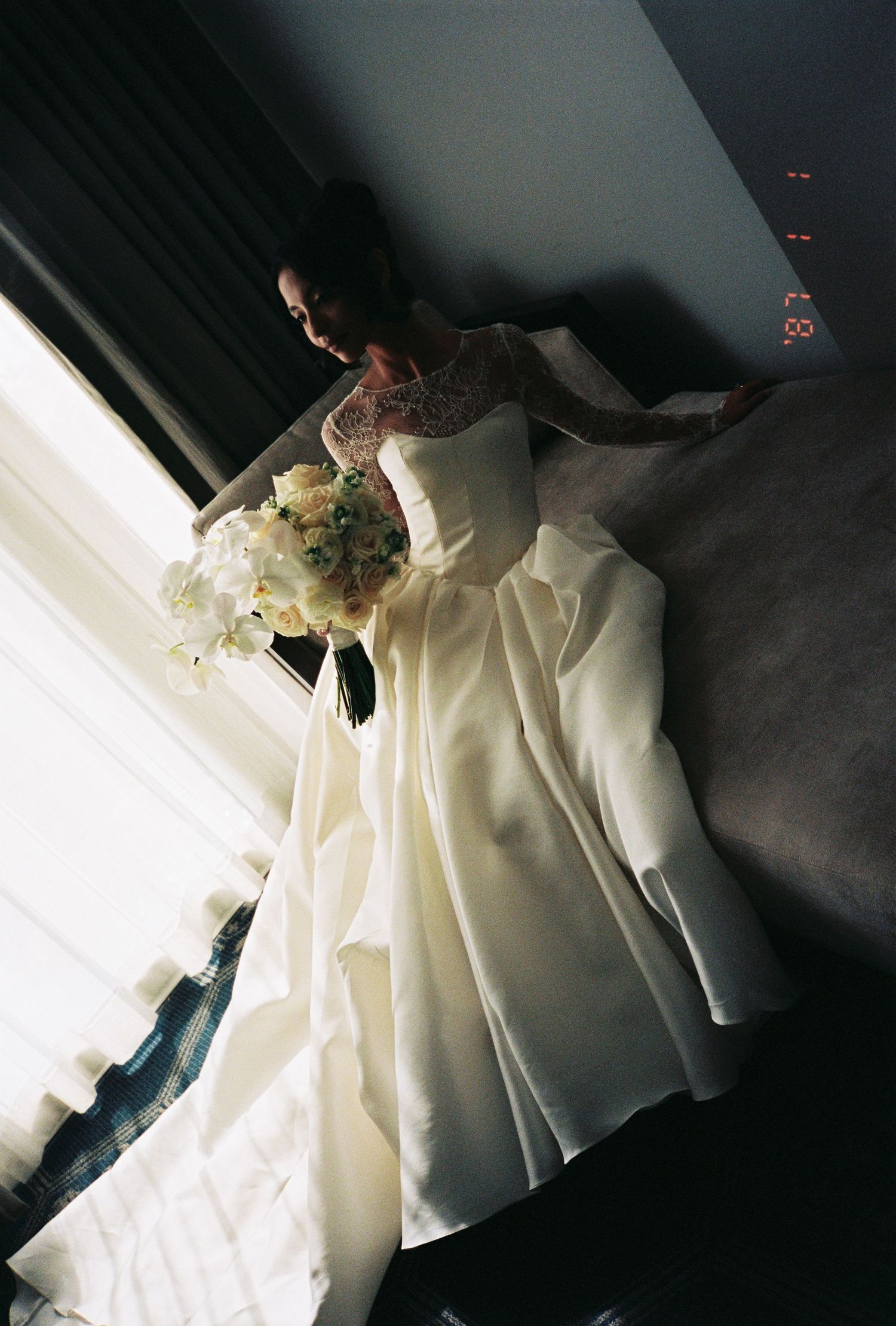 Bride in elegant white ball gown with lace sleeves holding a white floral bouquet near a window.