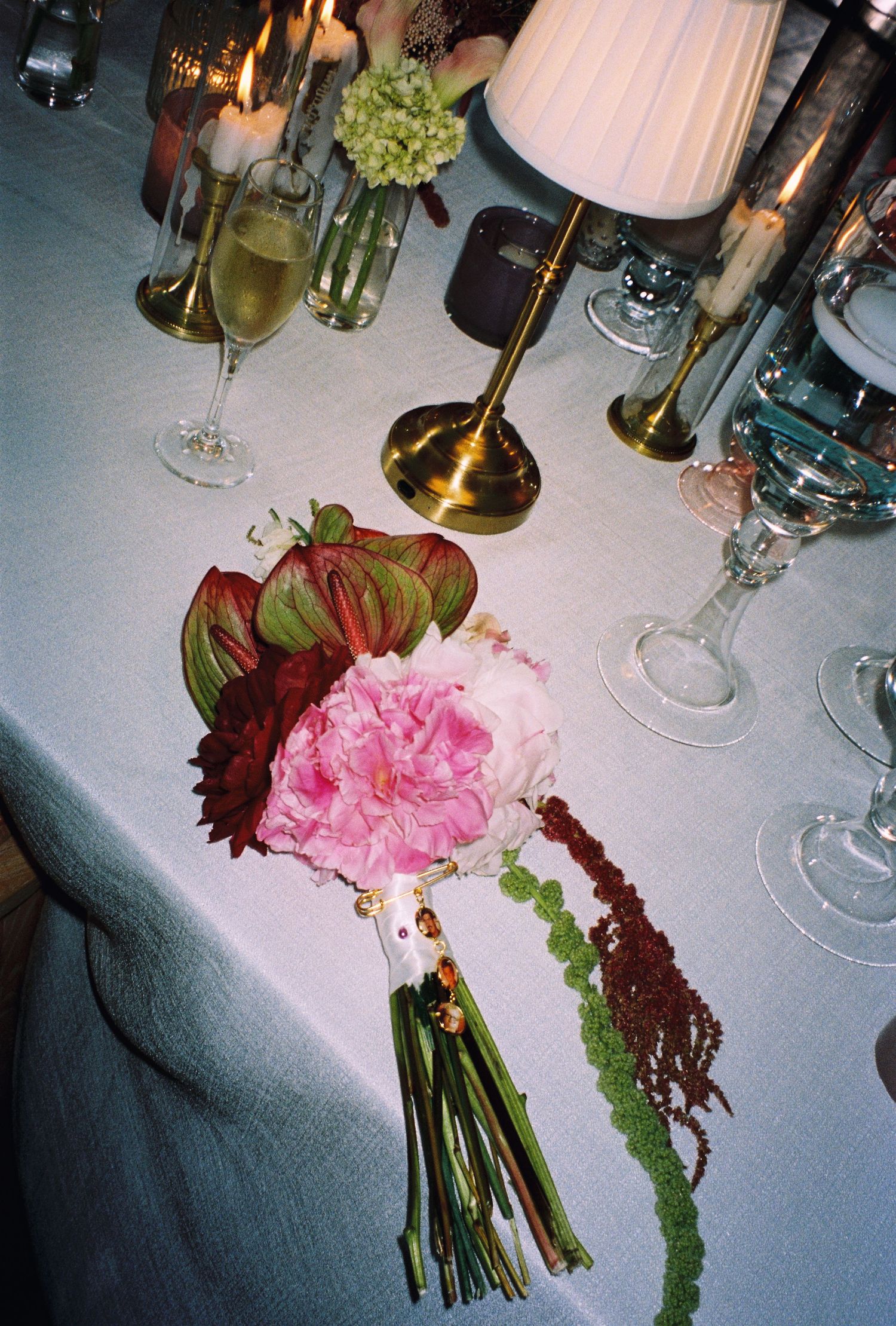 Pink peony and anthurium wedding bouquet on white tablecloth with champagne glasses and candles.