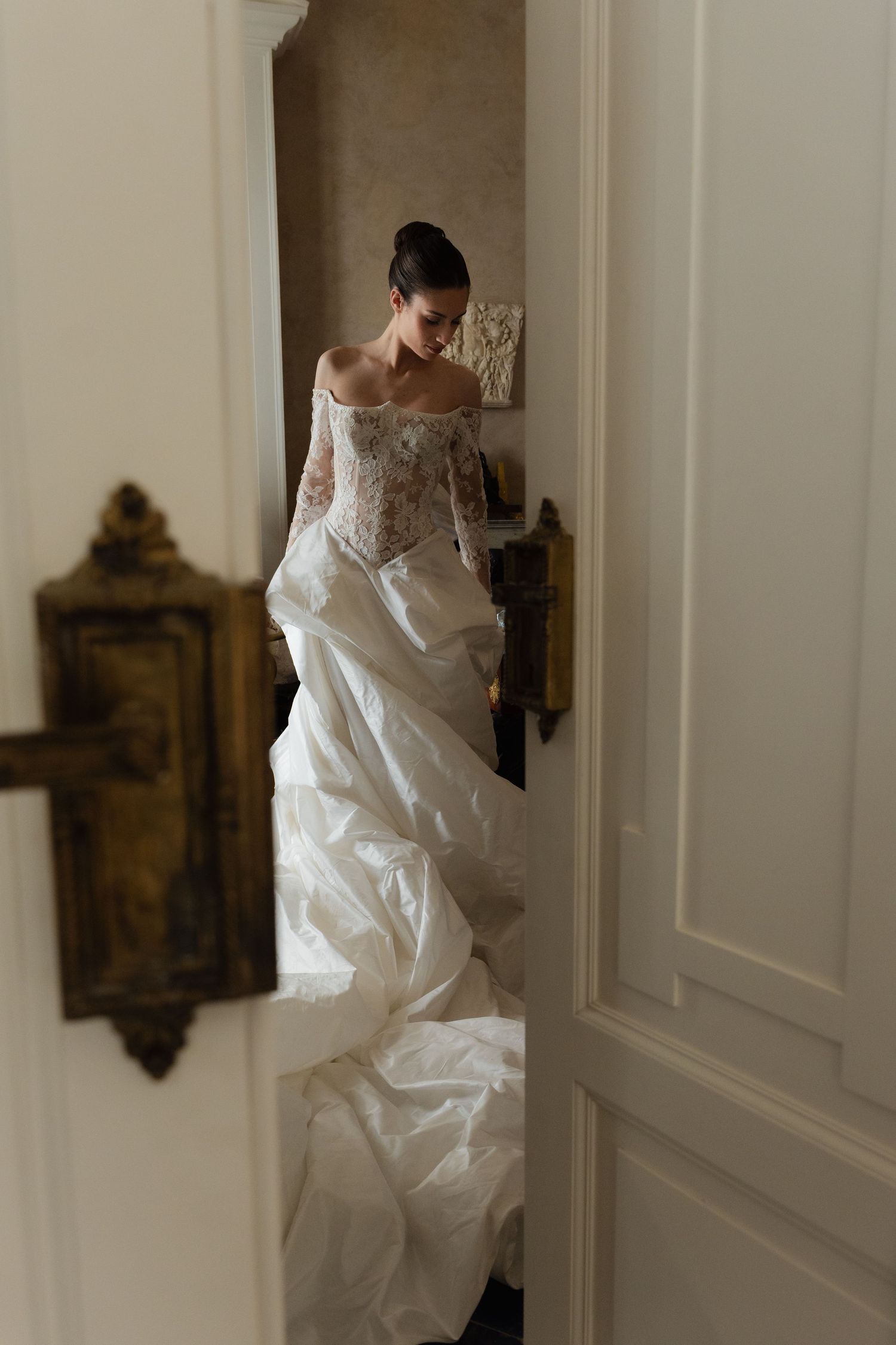 Bride in elegant off-shoulder lace wedding gown with voluminous skirt, standing in doorway of vintage room.