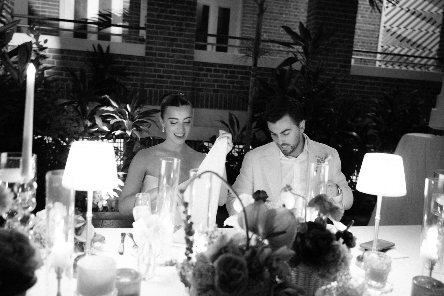 Black and white photo of a couple sharing a romantic dinner at an elegantly set table with candles and floral arrangements.