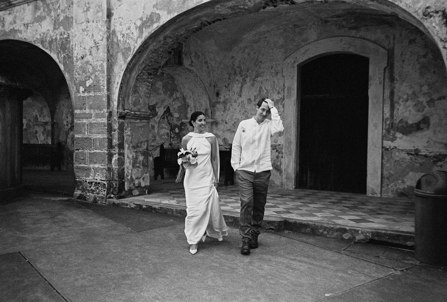 Black and white photo of a bride and groom walking through rustic stone archways at an outdoor wedding venue.