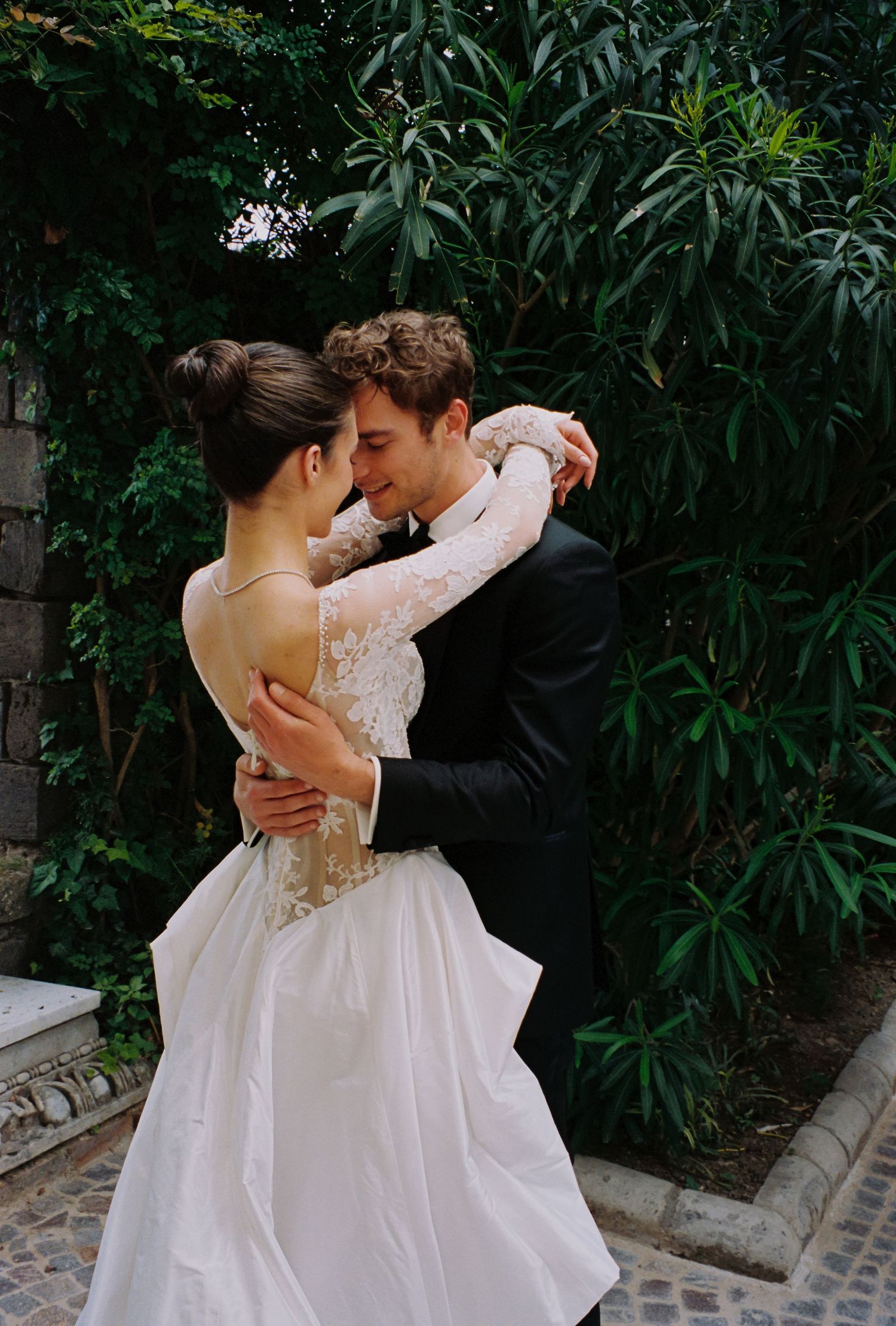 Bride and groom embracing outdoors, she wears a backless lace wedding gown, surrounded by lush green foliage.
