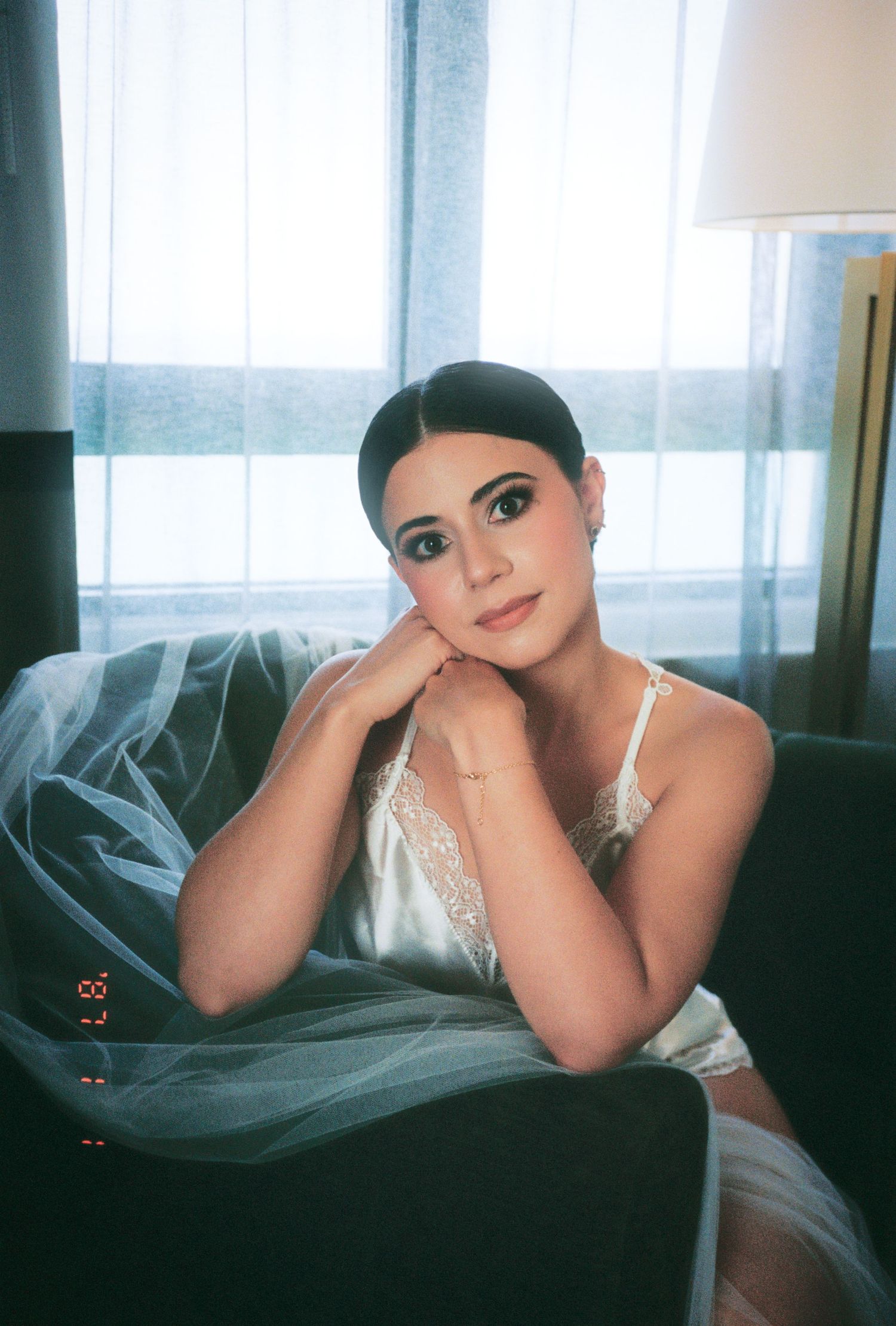 Elegant bride in white lace slip dress seated in chair with veil, wearing bridal makeup in hotel room.