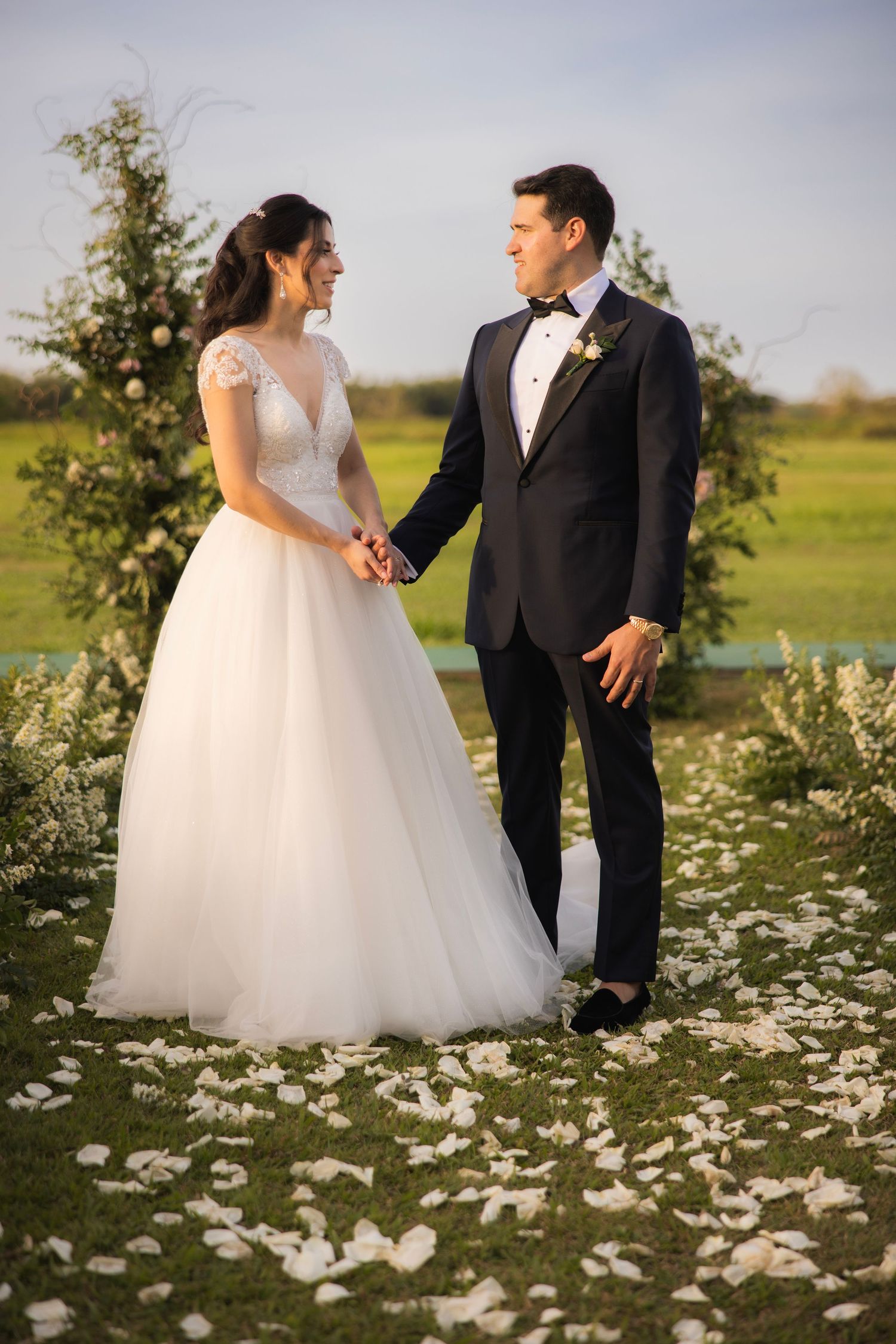 A couple in wedding attire poses on a grassy lawn decorated with white flower petals at sunset.