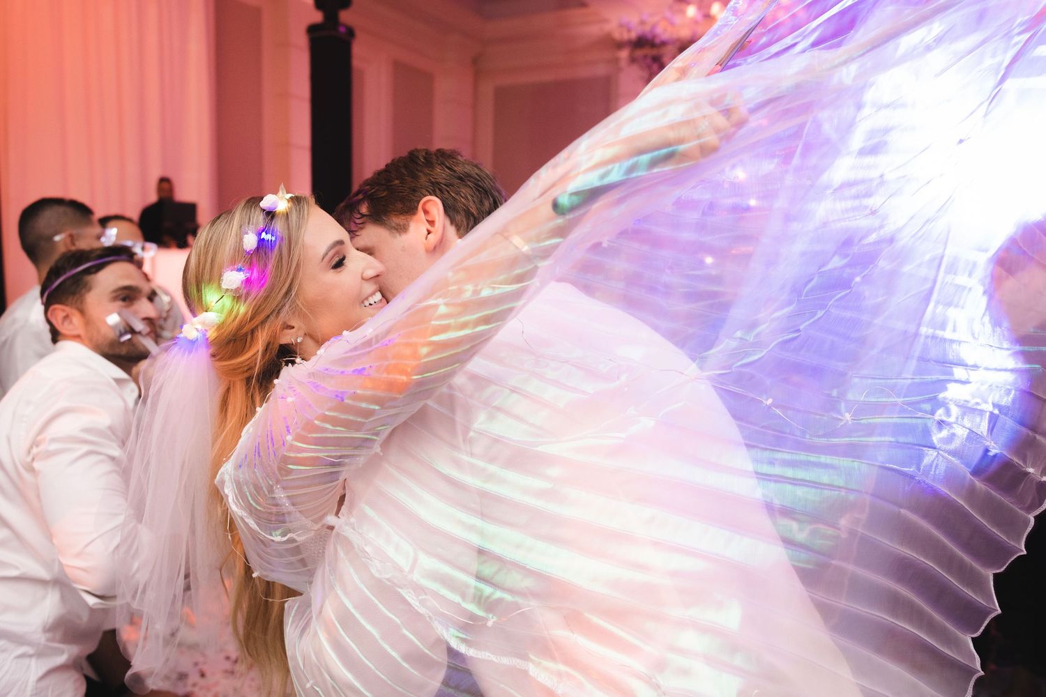 A couple shares a passionate dance moment under flowing iridescent fabric wings at their wedding celebration party.