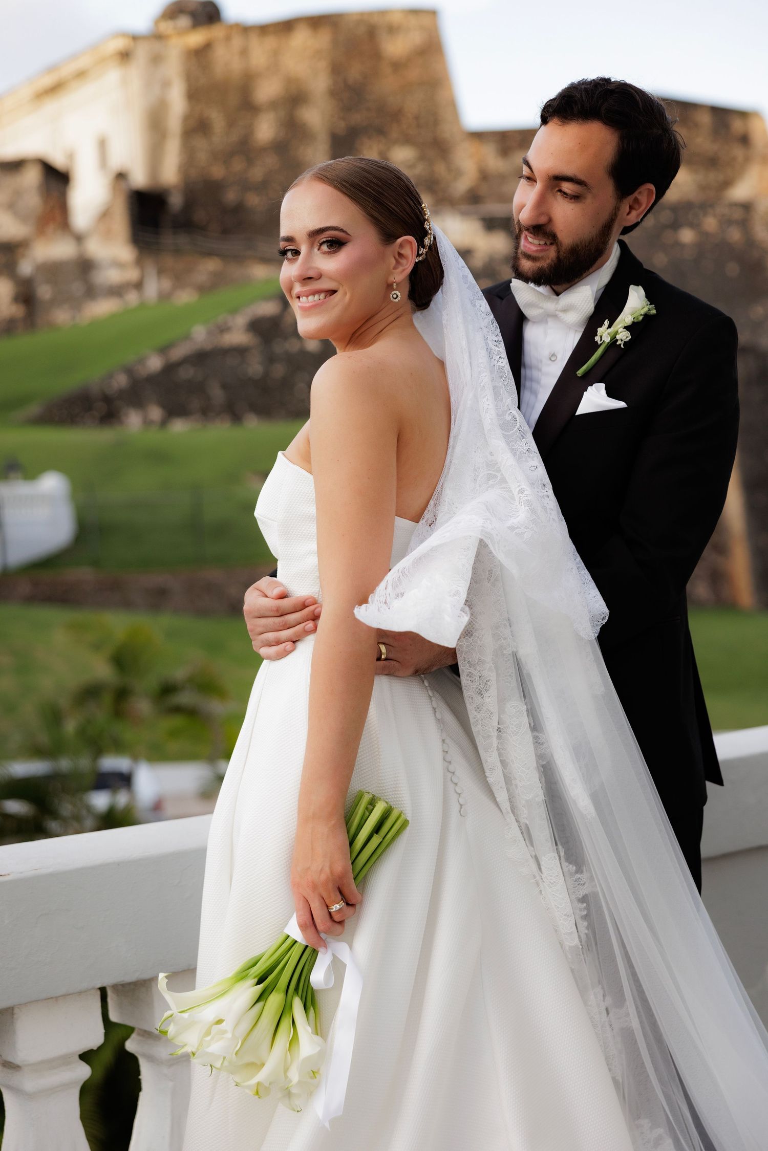 A wedding couple embraces on a terrace overlooking a historic stone fortress on their special day.