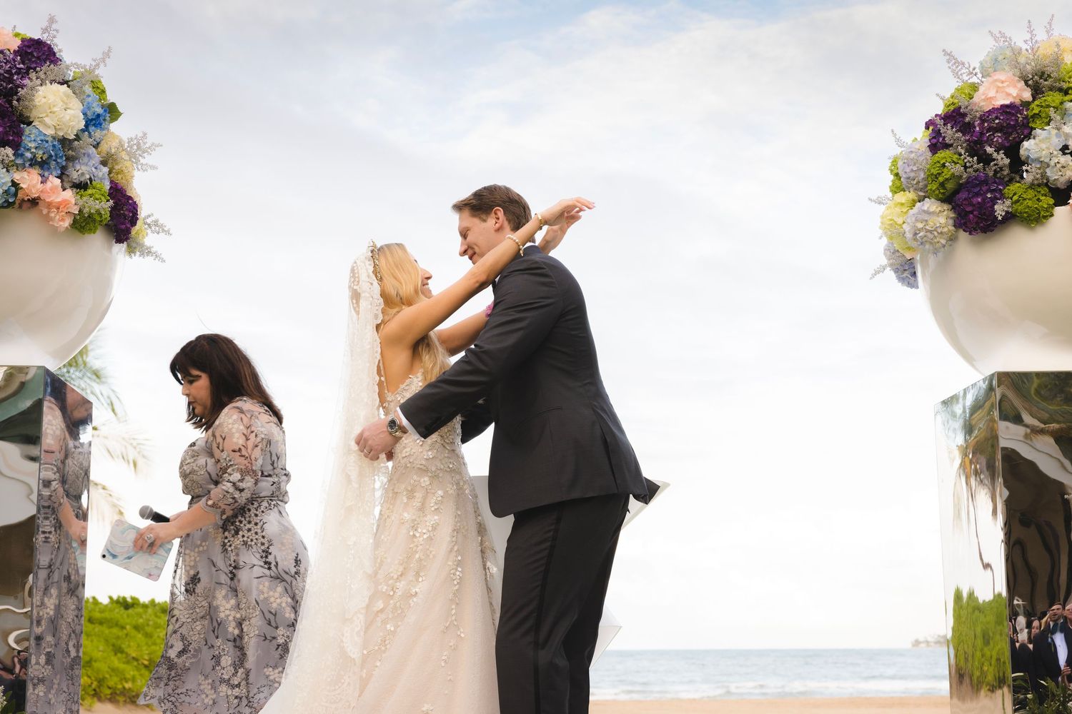 A romantic beach wedding ceremony with floral arrangements in white urns framing the couple as they embrace.