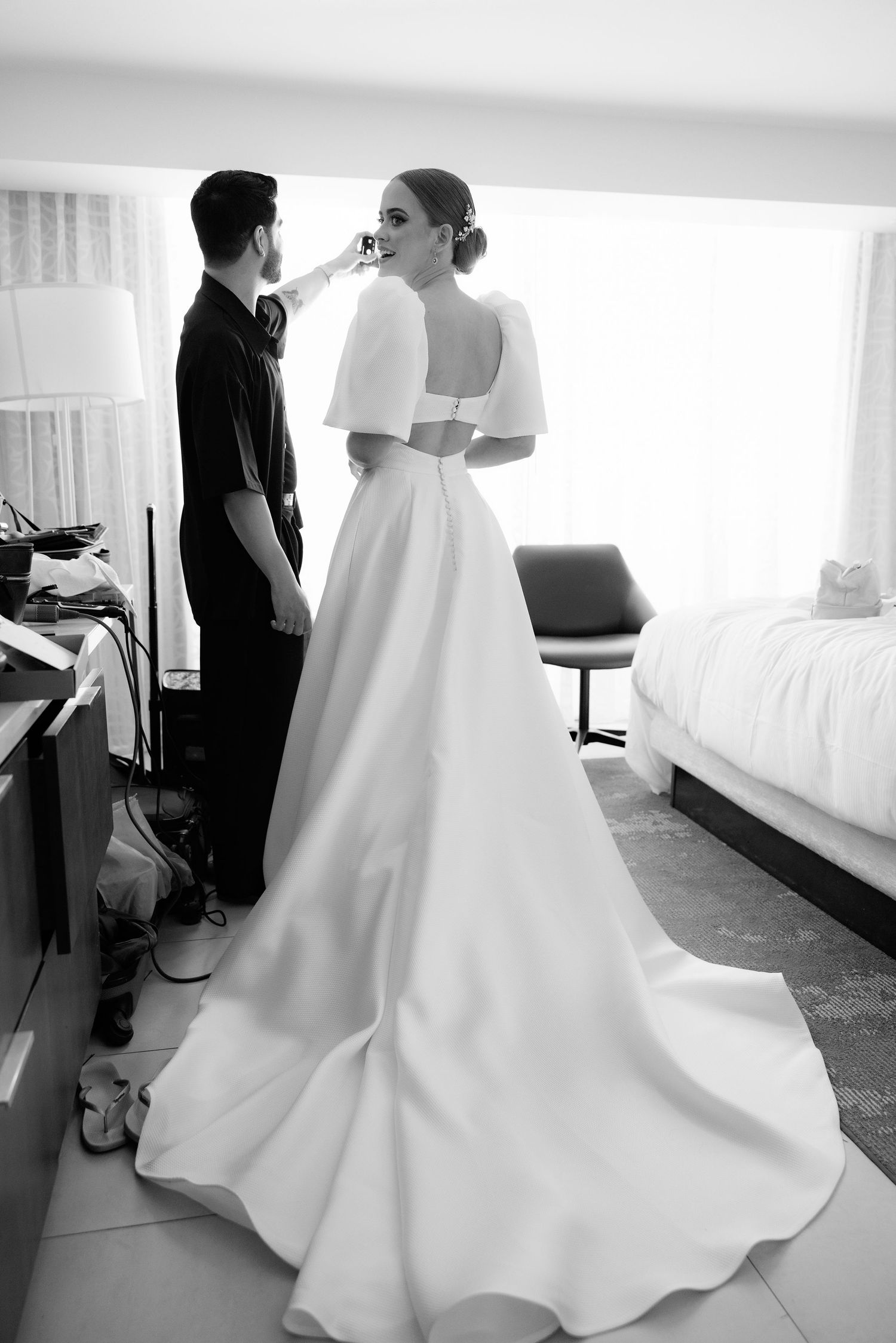Black and white photo of a couple getting ready before their wedding in a bright hotel room.