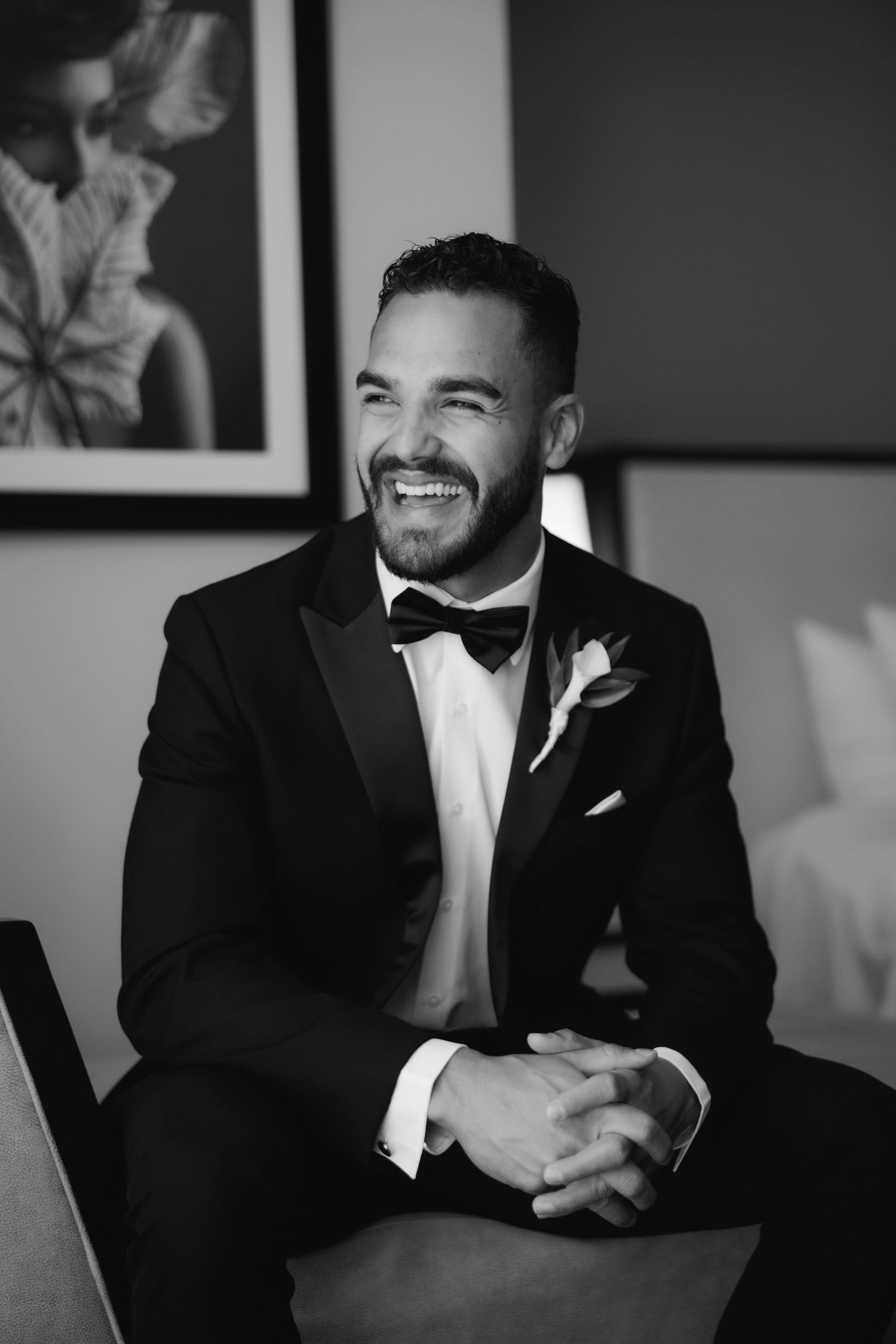 A joyful groom in a black tuxedo and bowtie sits and smiles during wedding preparations.