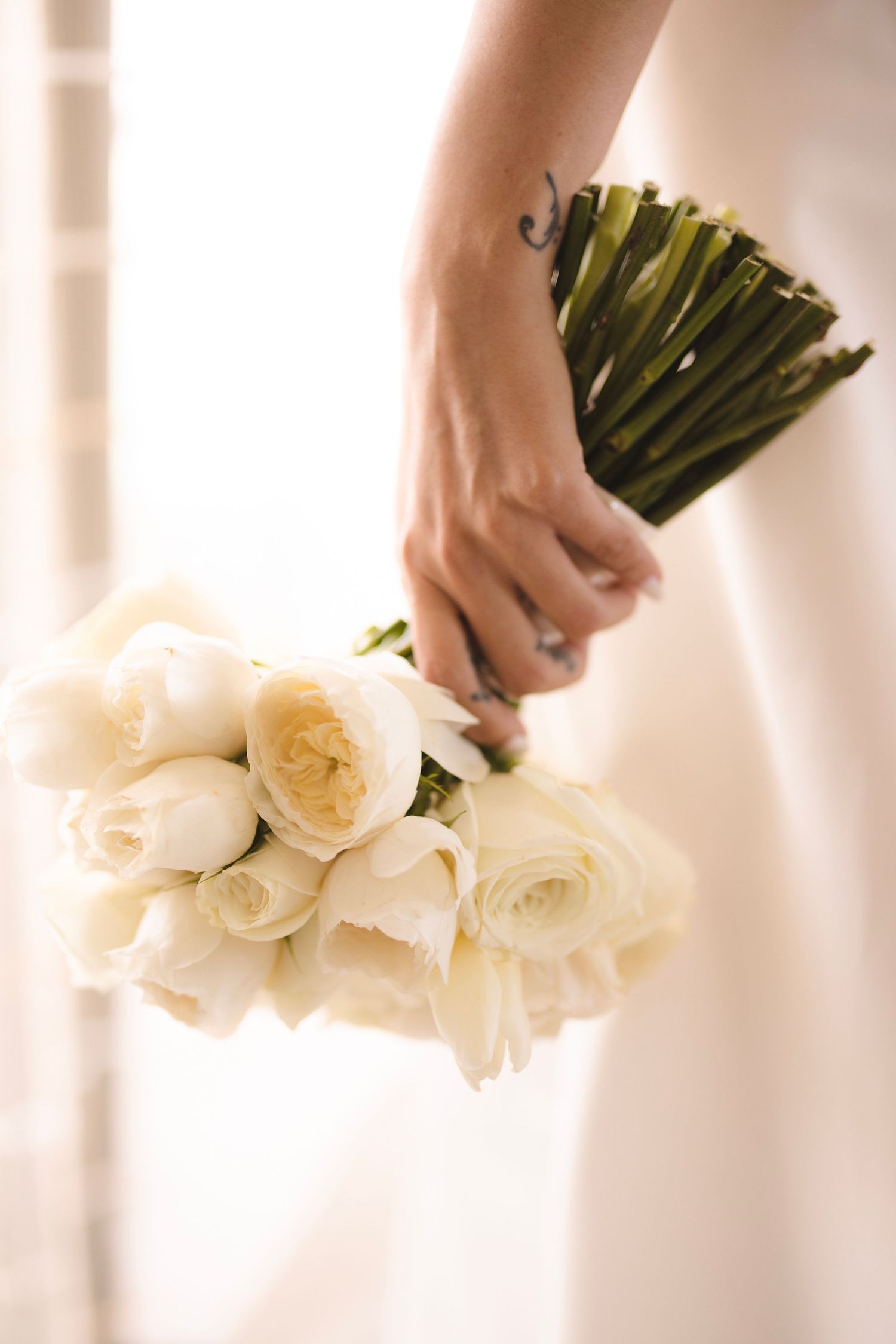 A hand gently holds a romantic bouquet of cream white roses wrapped with green stems against a soft light background.