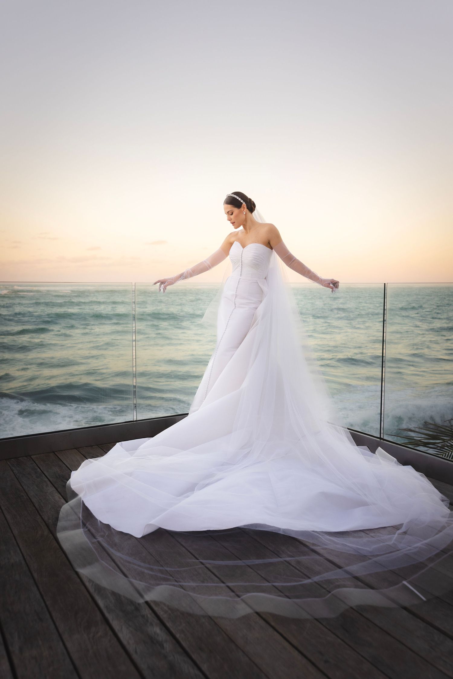 Flowing white wedding dress swirls gracefully on a wooden dock overlooking a serene ocean at sunset.