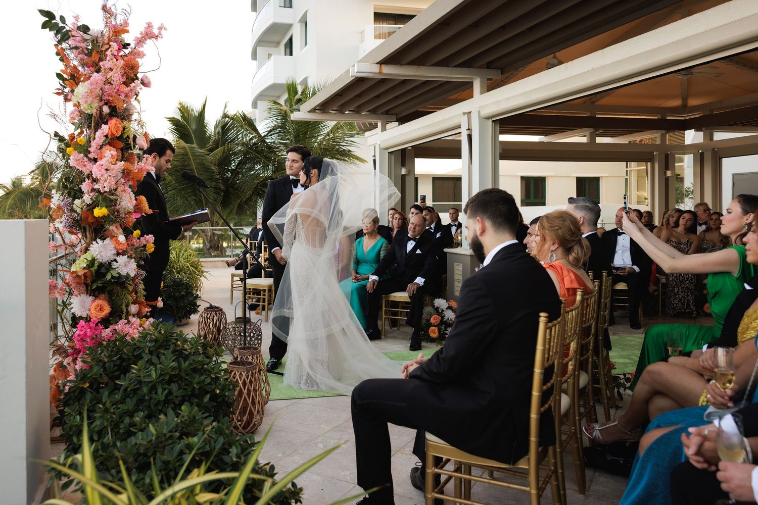 Wedding reception guests celebrate under open-air pavilion decorated with pink floral arrangements and string lights.