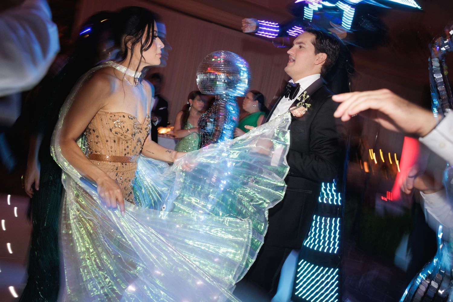 A couple dances energetically at a formal event with shimmering iridescent fabric catching the colorful lighting.